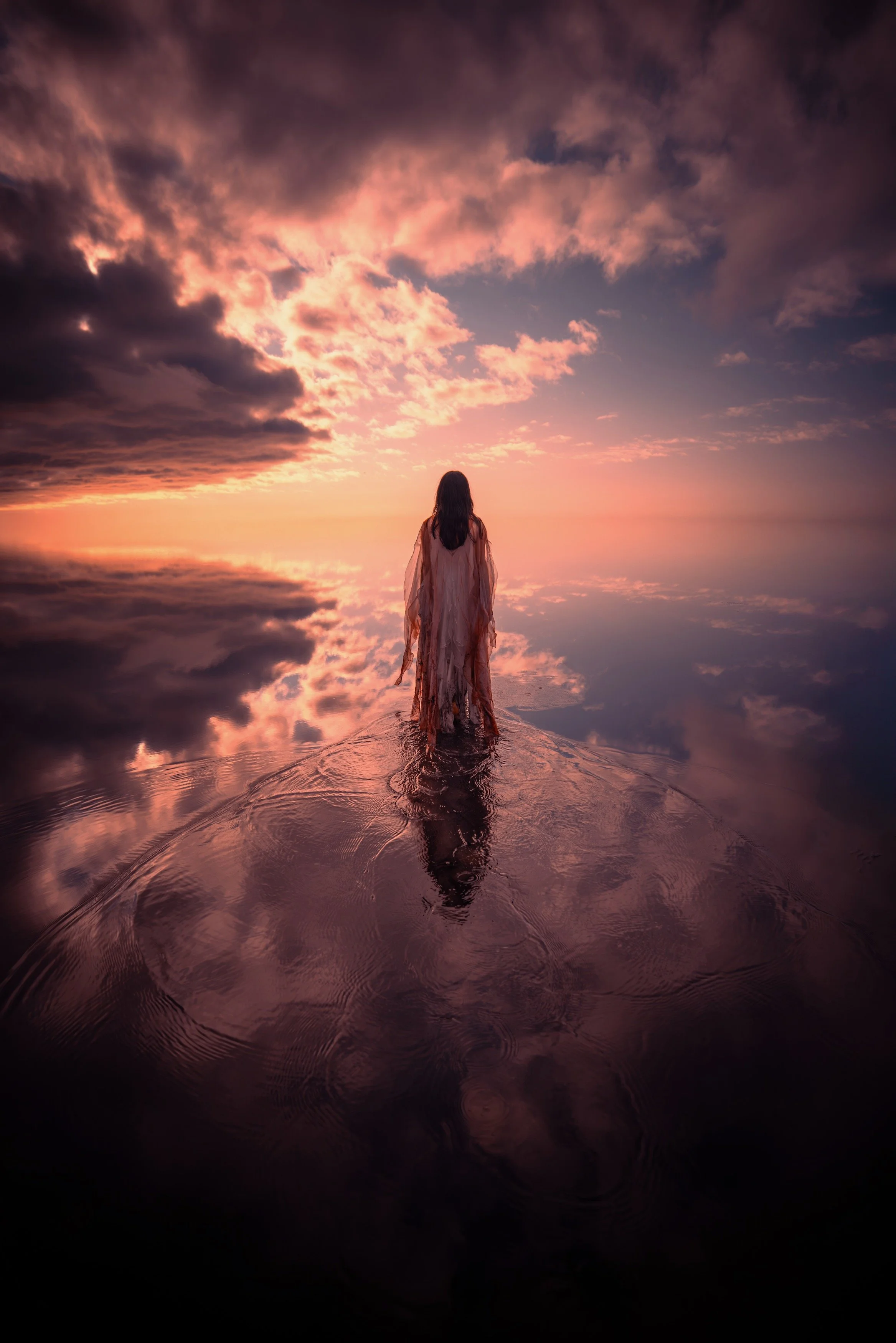 A woman in a flowing dress standing in shallow water at sunrise or sunset, with a colorful sky and clouds reflected on the water's surface.