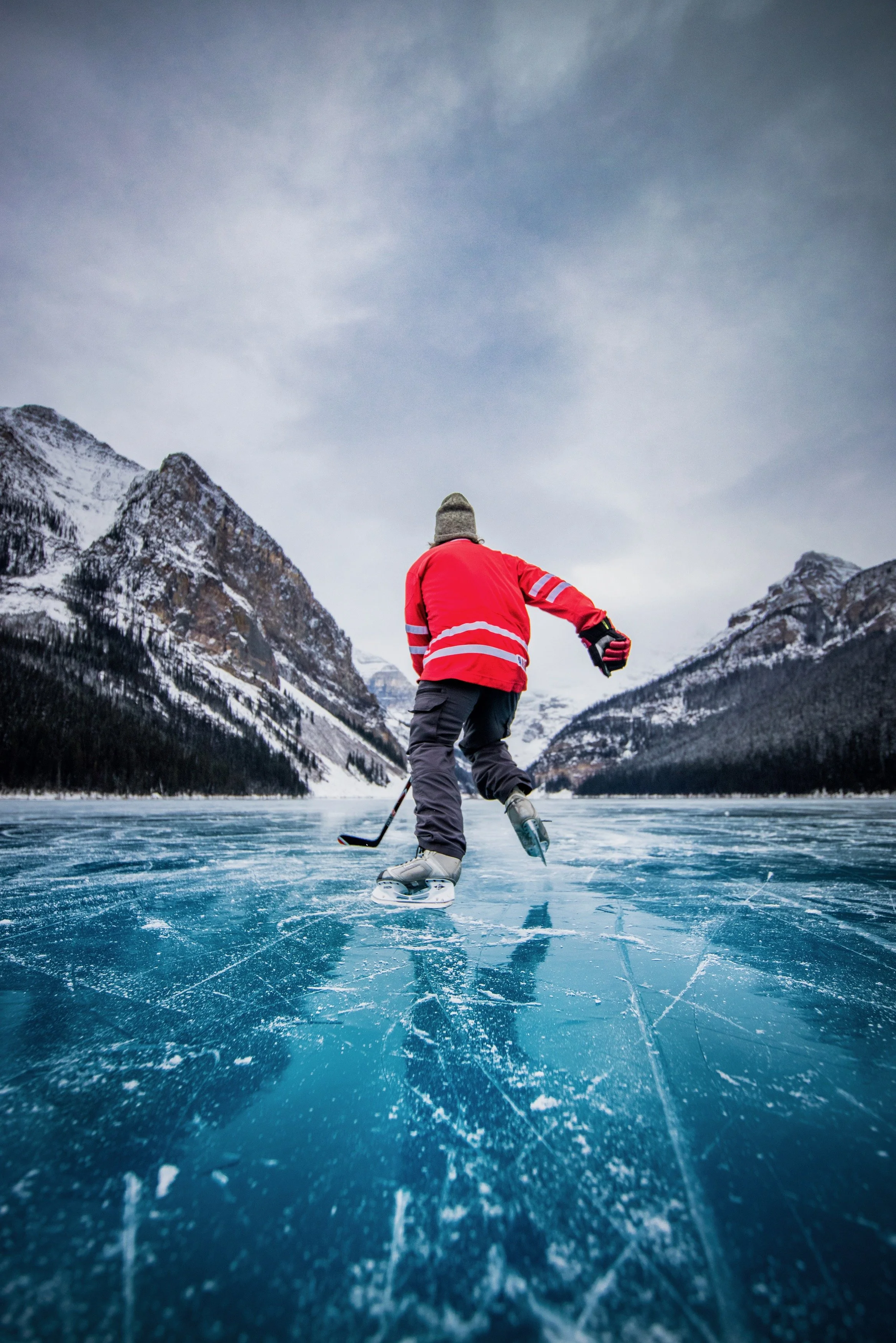 Person dressed in red hockey jersey, gray beanie, and black pants ice skating on a frozen Lake Louise in Banff National Park, Canada, surrounded by snow-capped mountains.