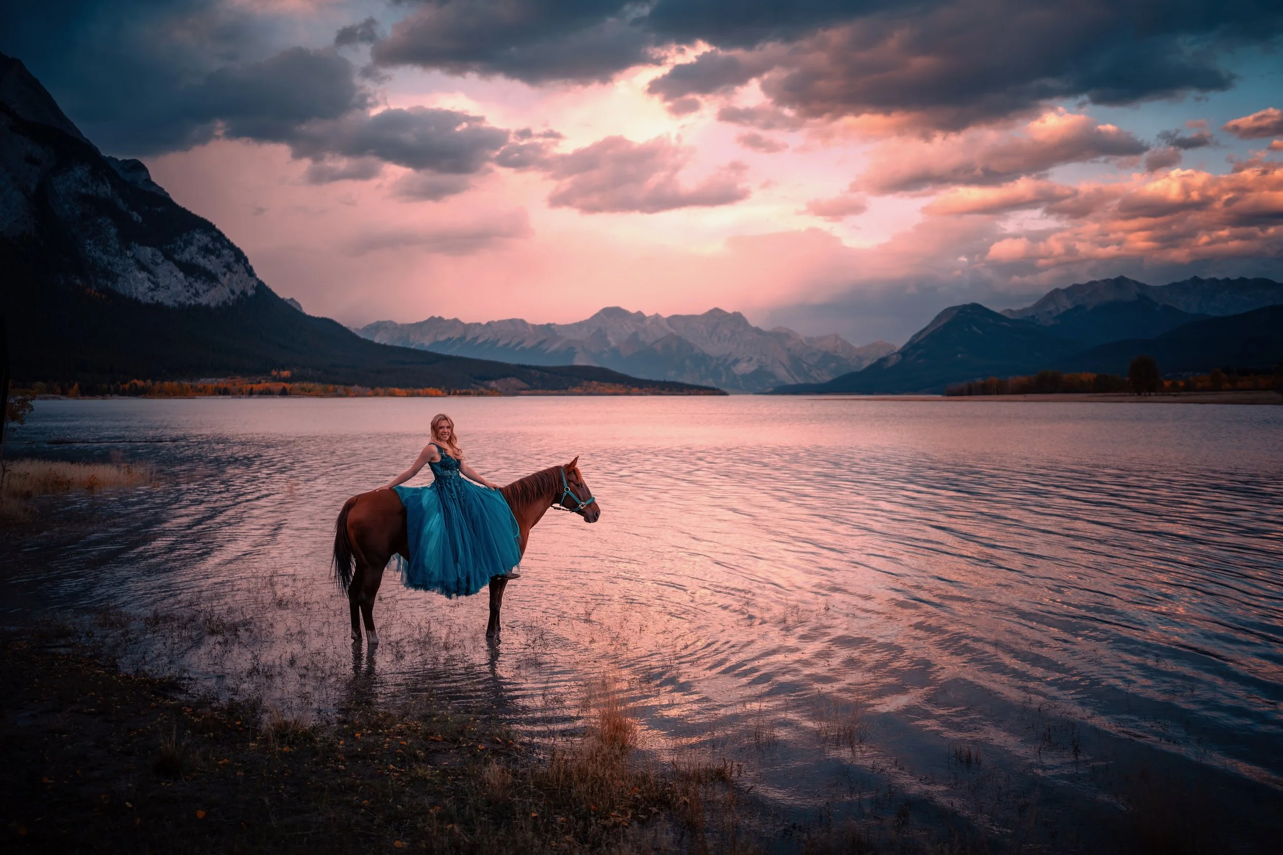 A woman in a blue dress sitting on a brown horse standing in a calm lake during sunset, with mountains in the background and a partly cloudy sky.