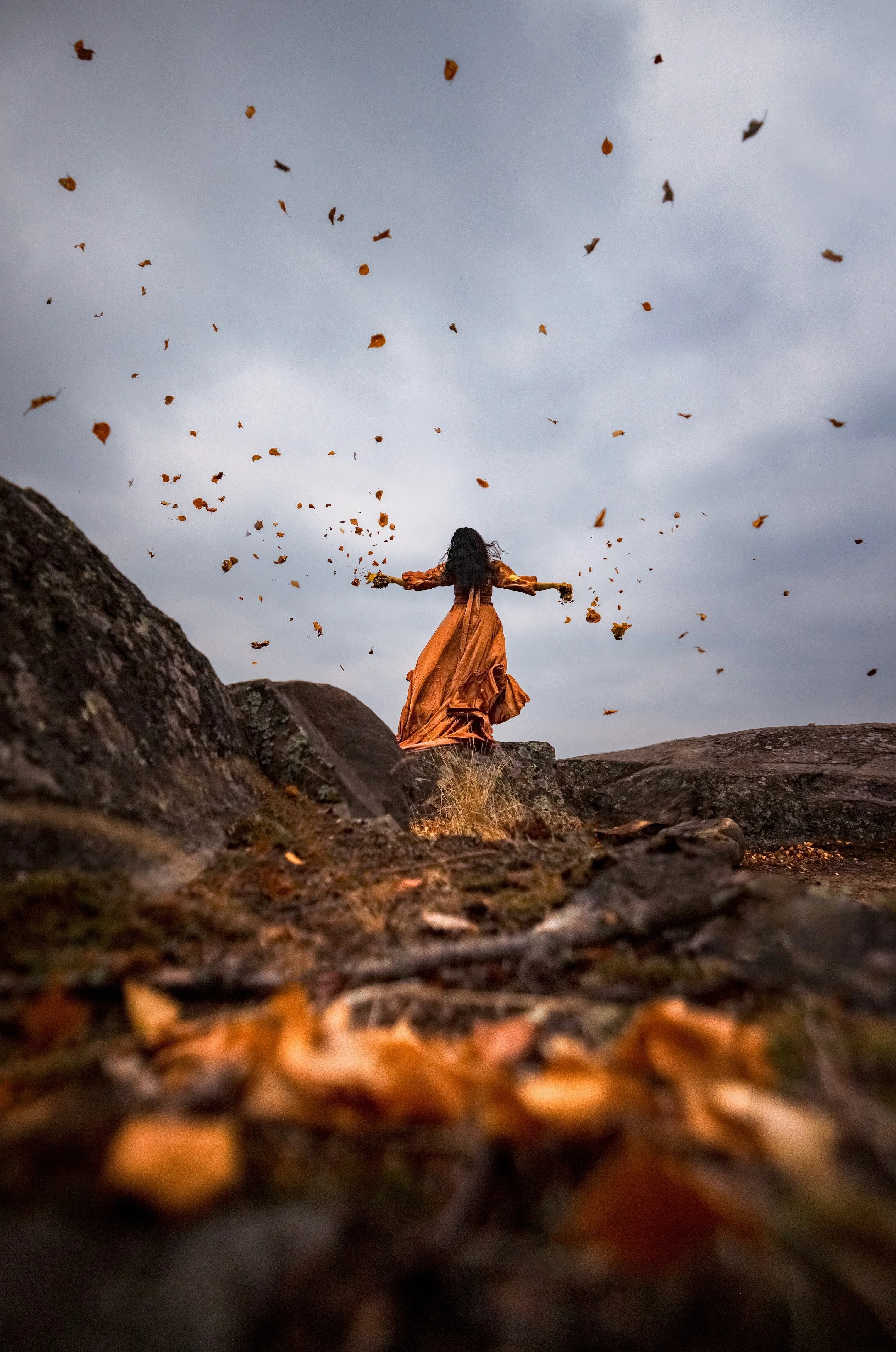 A woman in an orange dress standing on rocks with leaves falling around her under a cloudy sky.