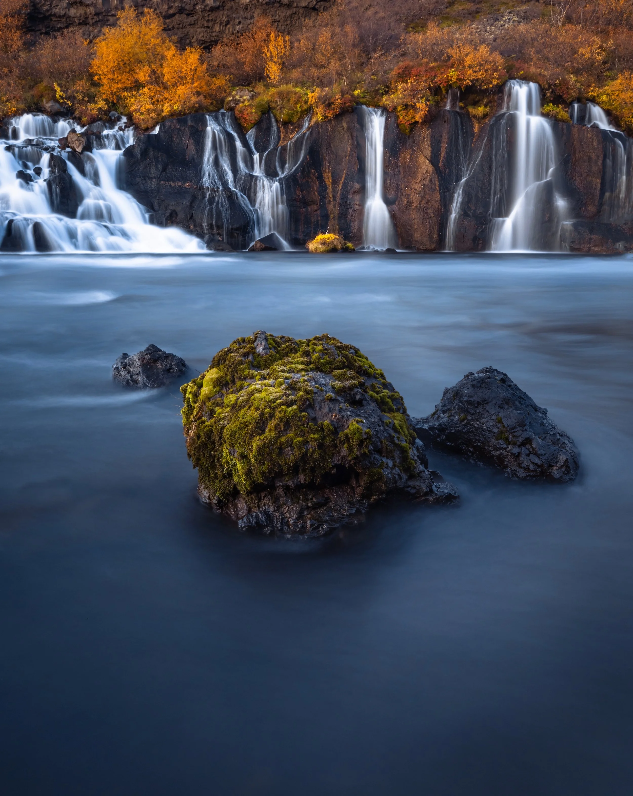A waterfall cascading down a rocky cliff into a river with moss-covered rocks in the foreground, surrounded by autumn foliage.