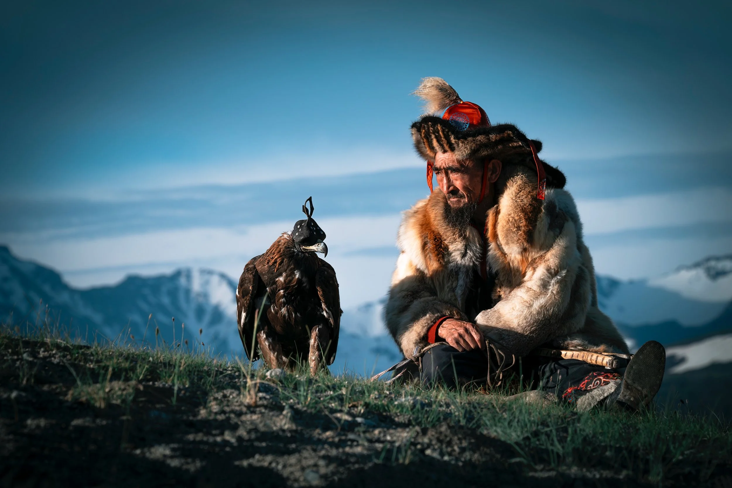 Mongolian man in traditional clothing sitting outdoors with a golden eagle and mountain landscape in background.