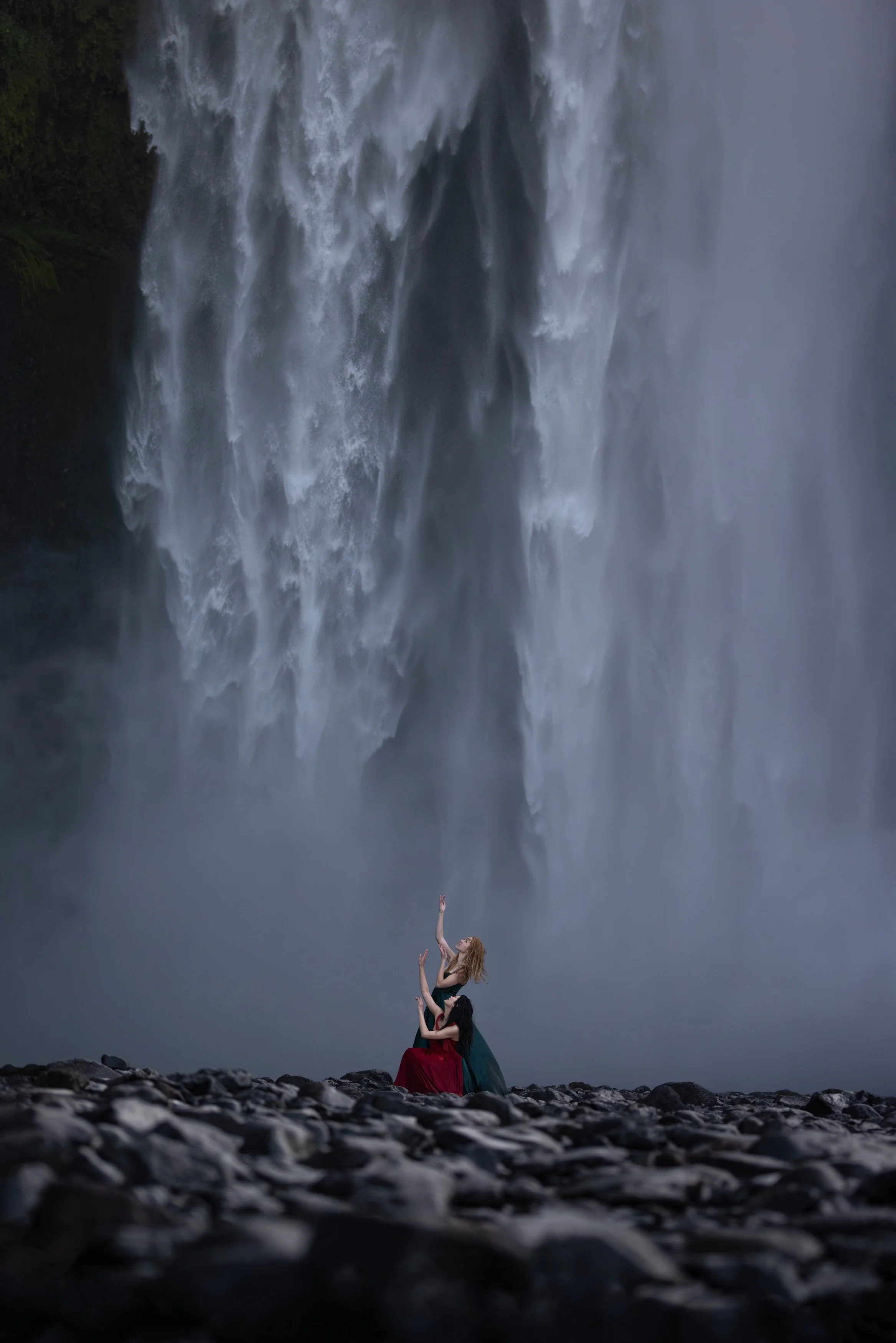 Two women in elegant dresses dancing on rocky ground in front of a large waterfall.
