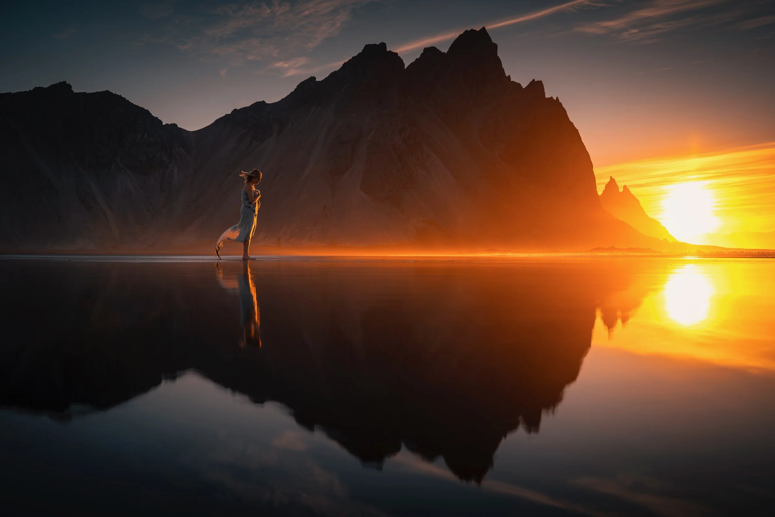 A person walking on a reflective wet surface at sunset with mountains in the background and a vibrant orange and yellow sky.