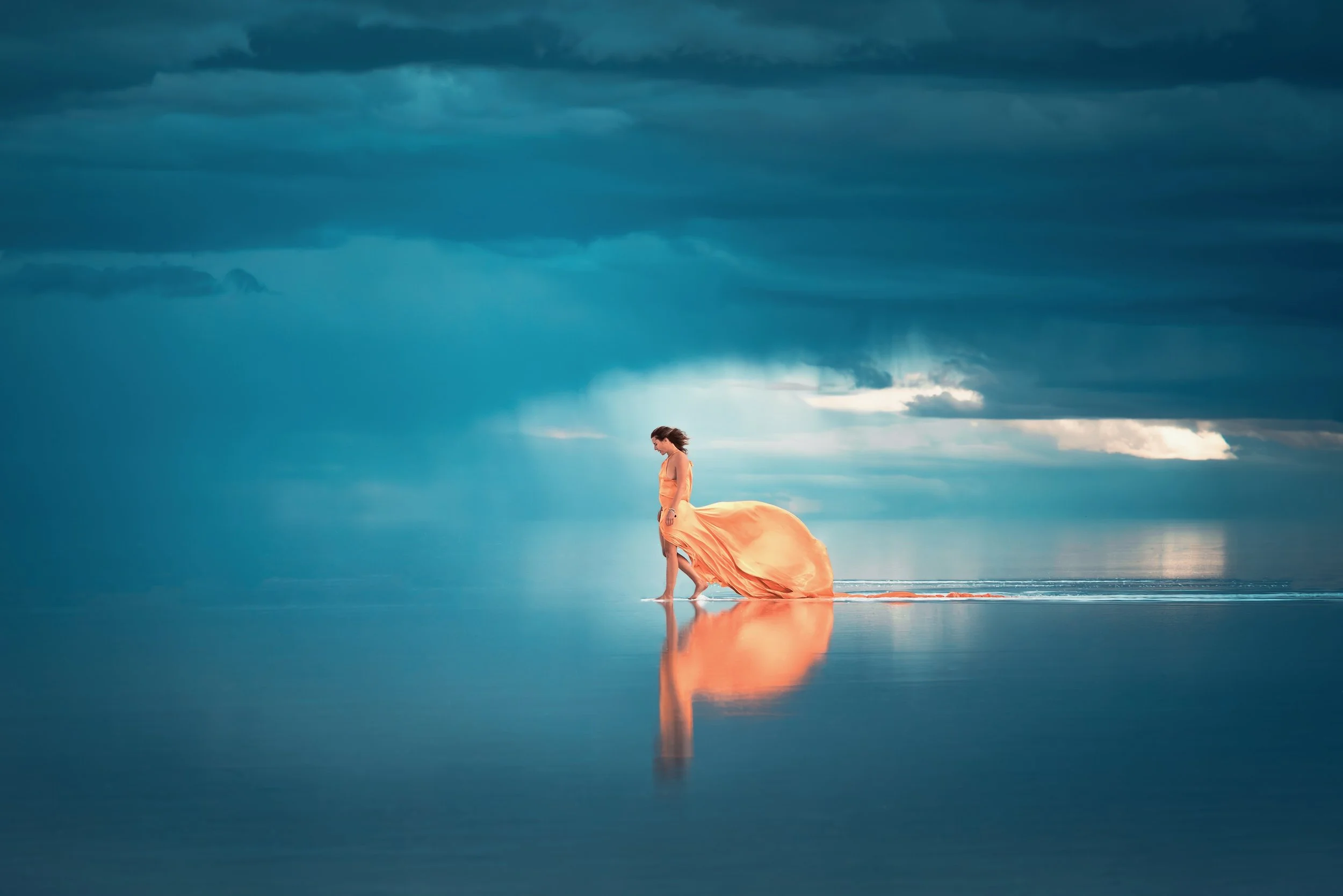 A woman in a flowing peach-colored dress walking barefoot on wet sand near the ocean, with a dramatic cloudy sky in the background and her reflection visible on the sand.