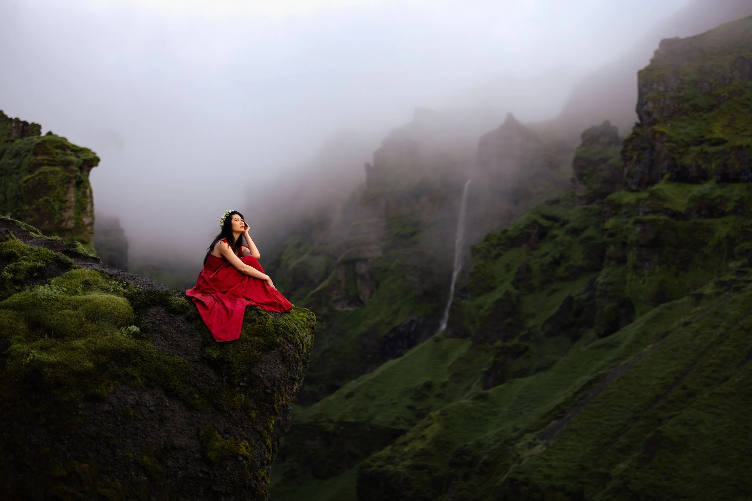 A woman in a red dress sitting on a moss-covered rocky ledge in a misty mountain landscape with green slopes and waterfalls in the background.