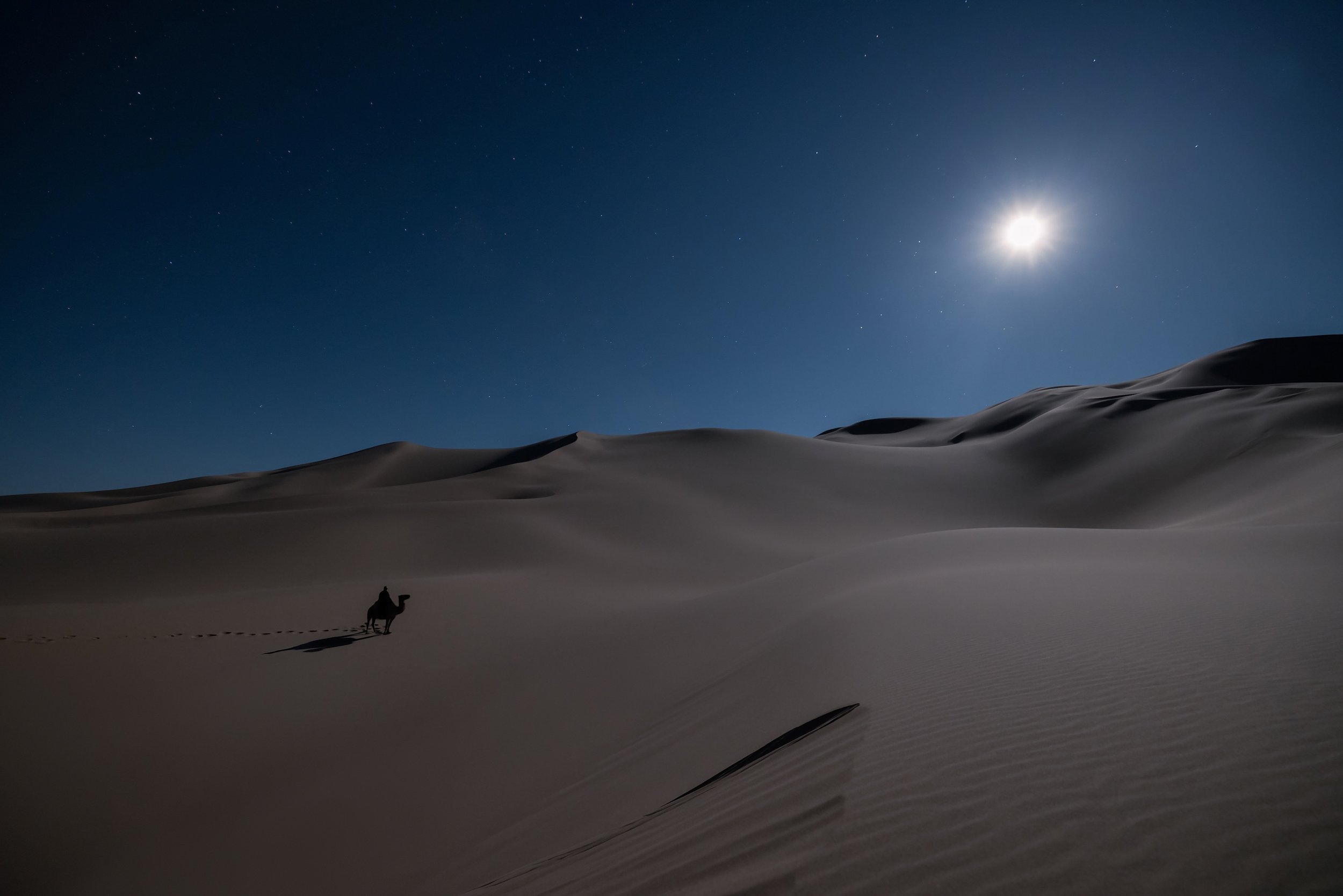 A person riding a camel across sandy desert dunes under a night sky with a bright moon and stars.
