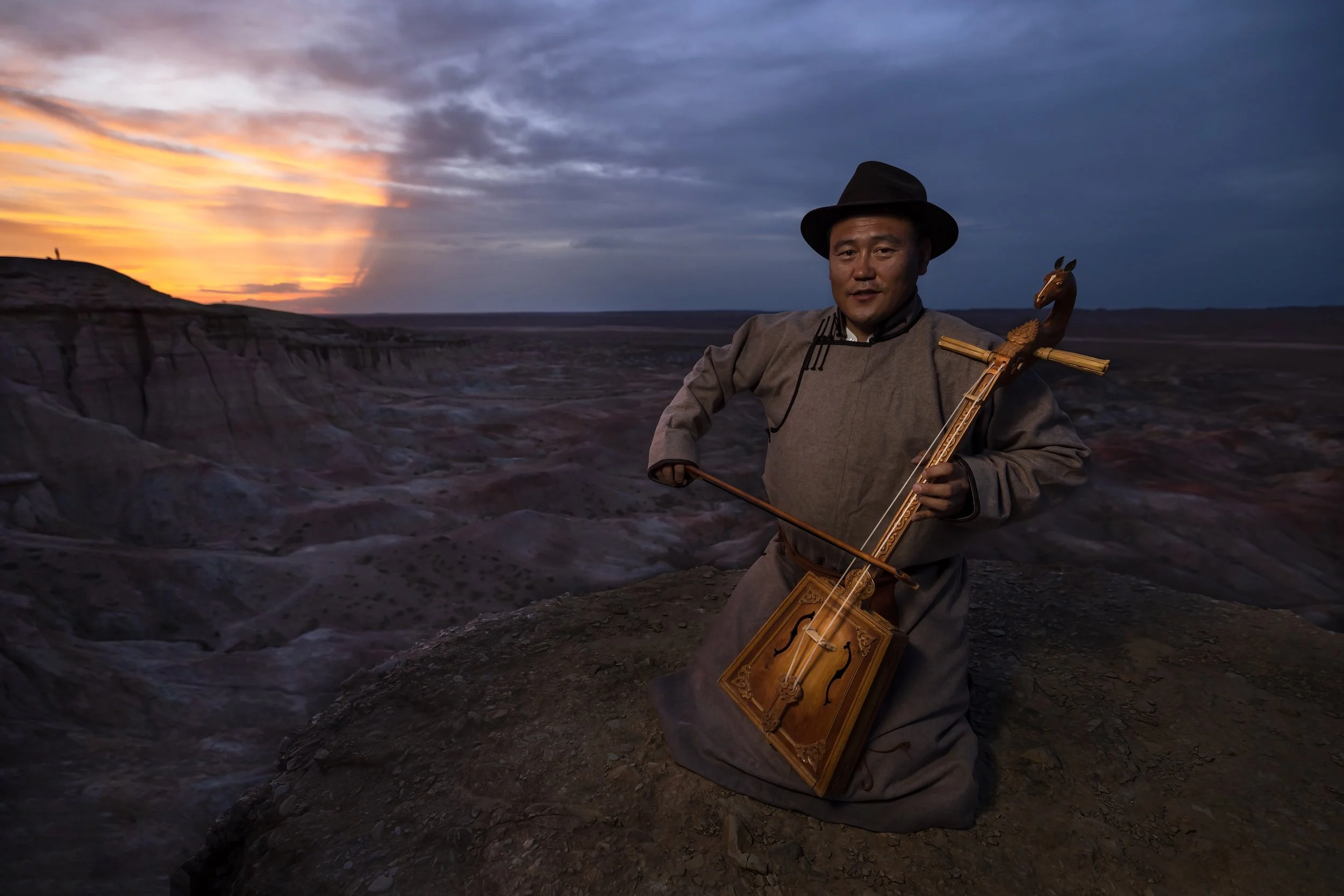A man dressed in traditional clothing kneeling on a rocky ground, playing a bowed string instrument resembling a morin khuur, with a desert landscape and sunset sky in the background.