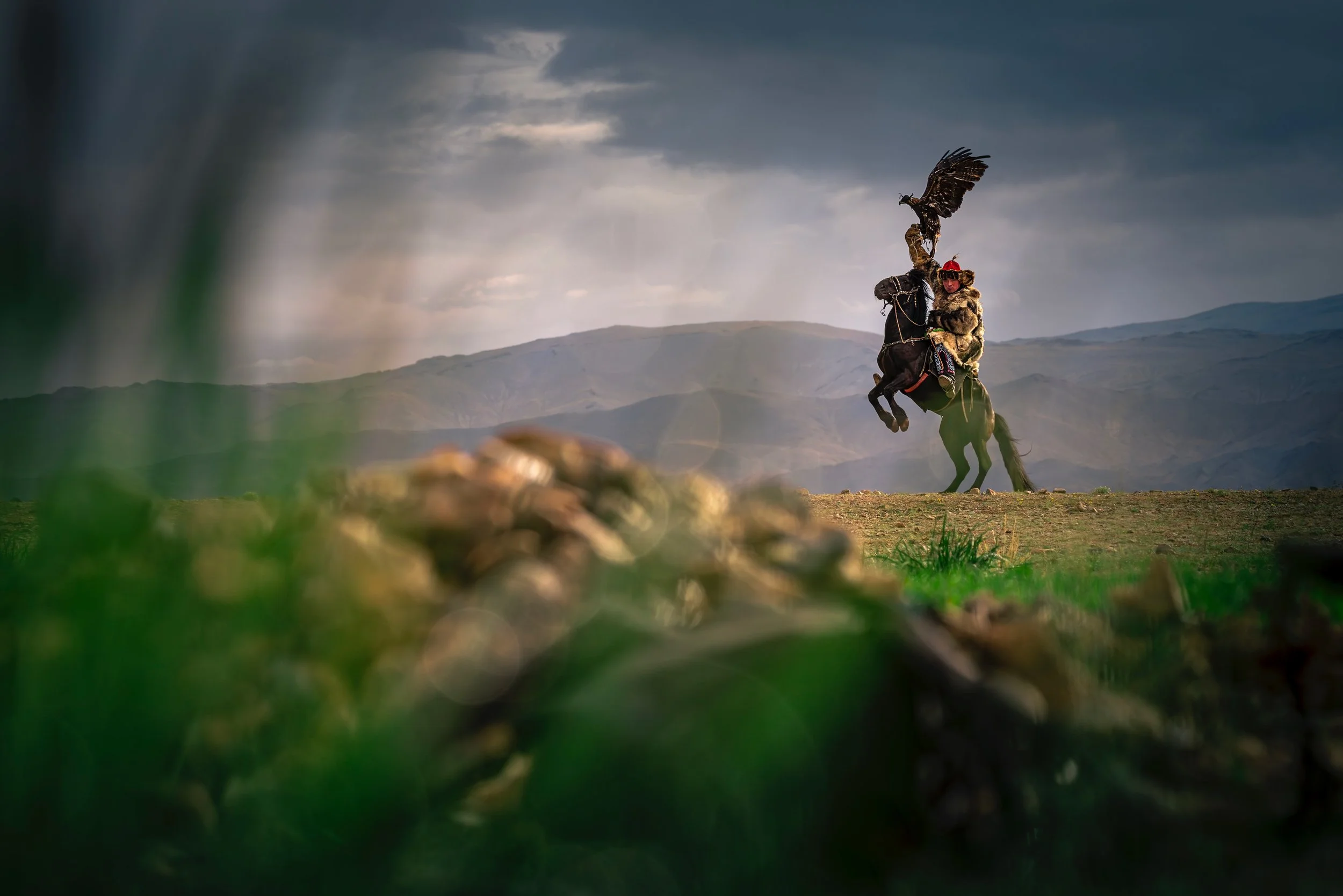 A performer riding a rearing horse with a large bird on their arm, set against a mountainous landscape under a cloudy sky.