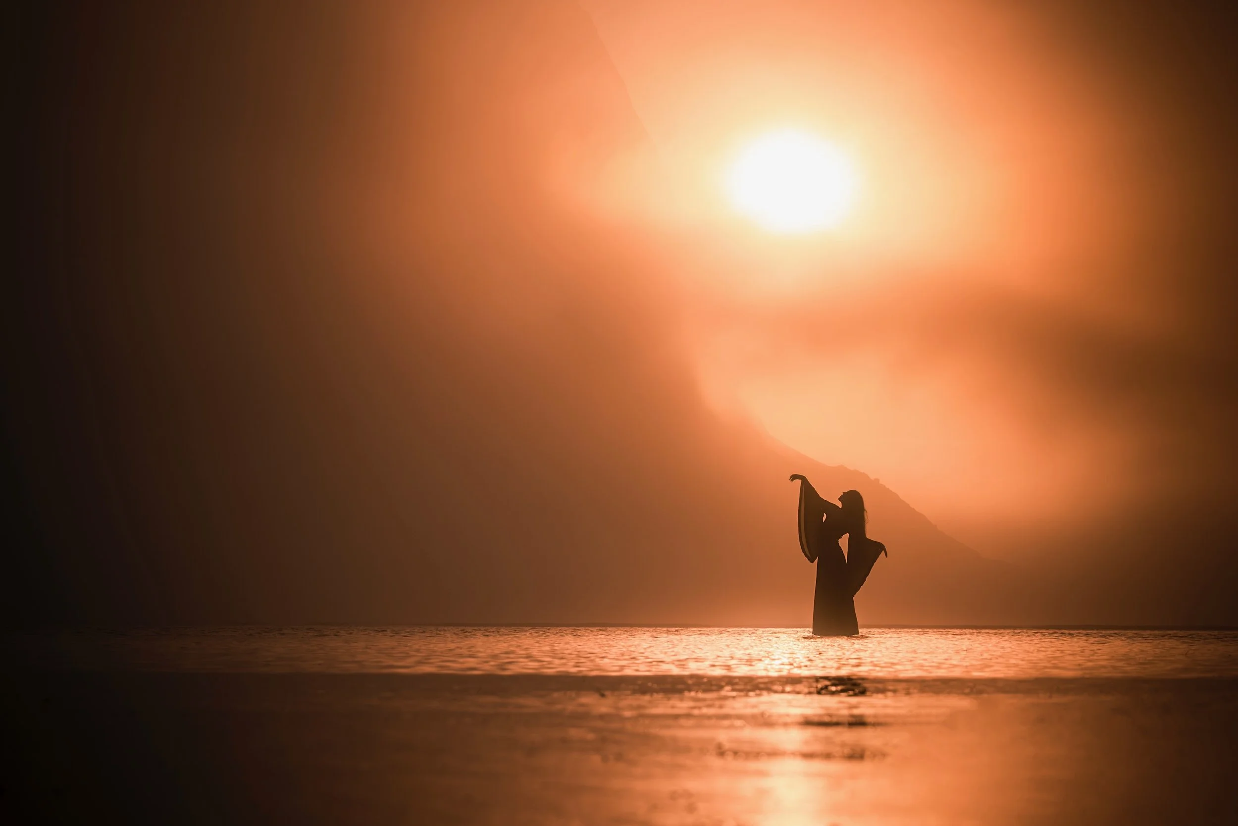 Silhouette of a woman in the water at sunset, holding a surfboard, with a mountainous landscape in the background.