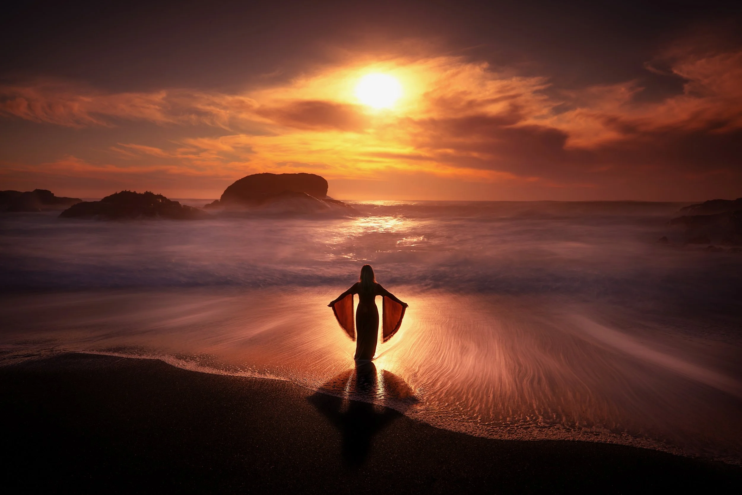 Silhouette of a woman standing on a beach at sunset, facing the ocean with arms outstretched, wearing a flowing dress, with rocks and the sun setting over the horizon.