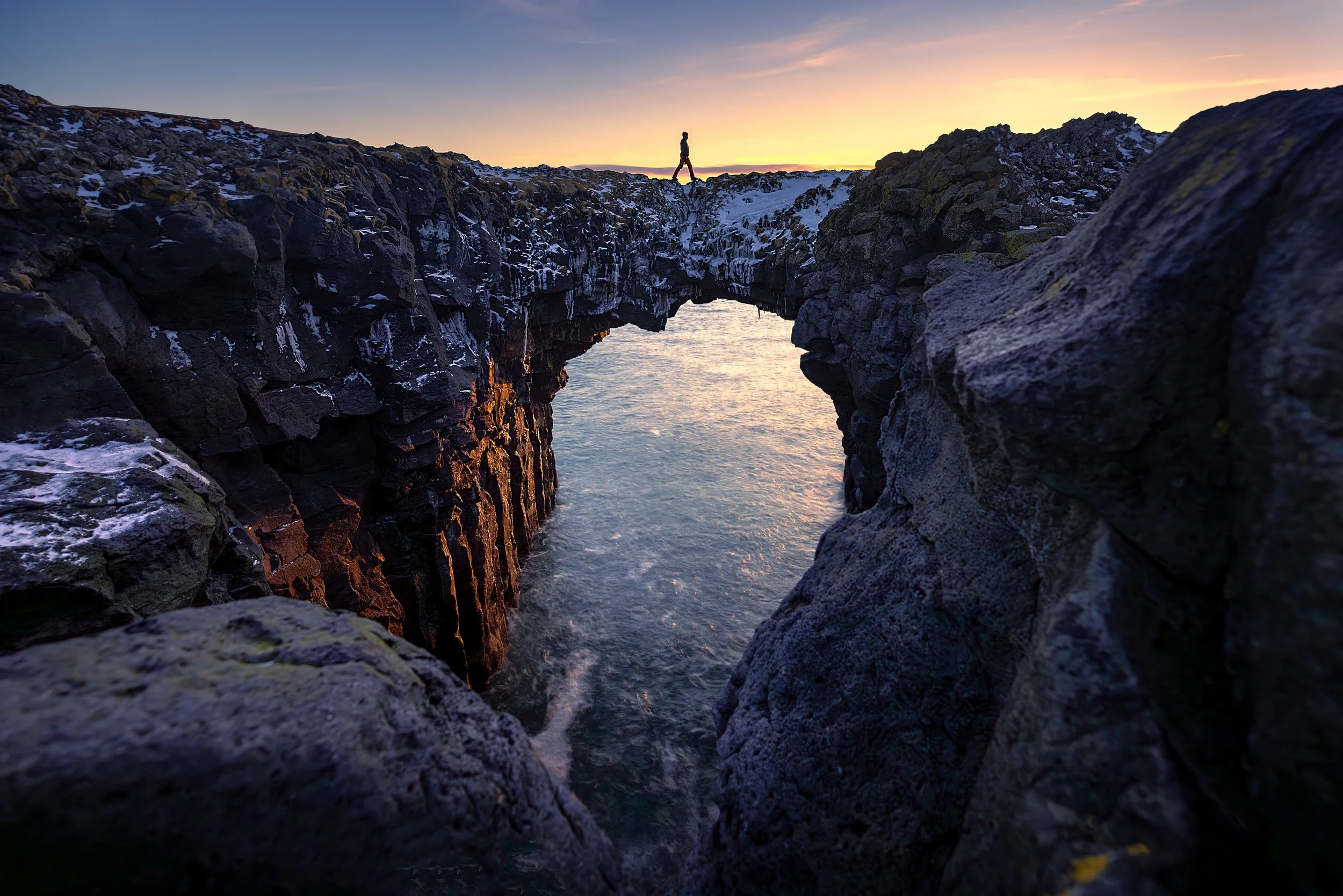 A person walking on top of a rock formation with a natural arch over a water body during sunset, with snow on the ground and a colorful sky.