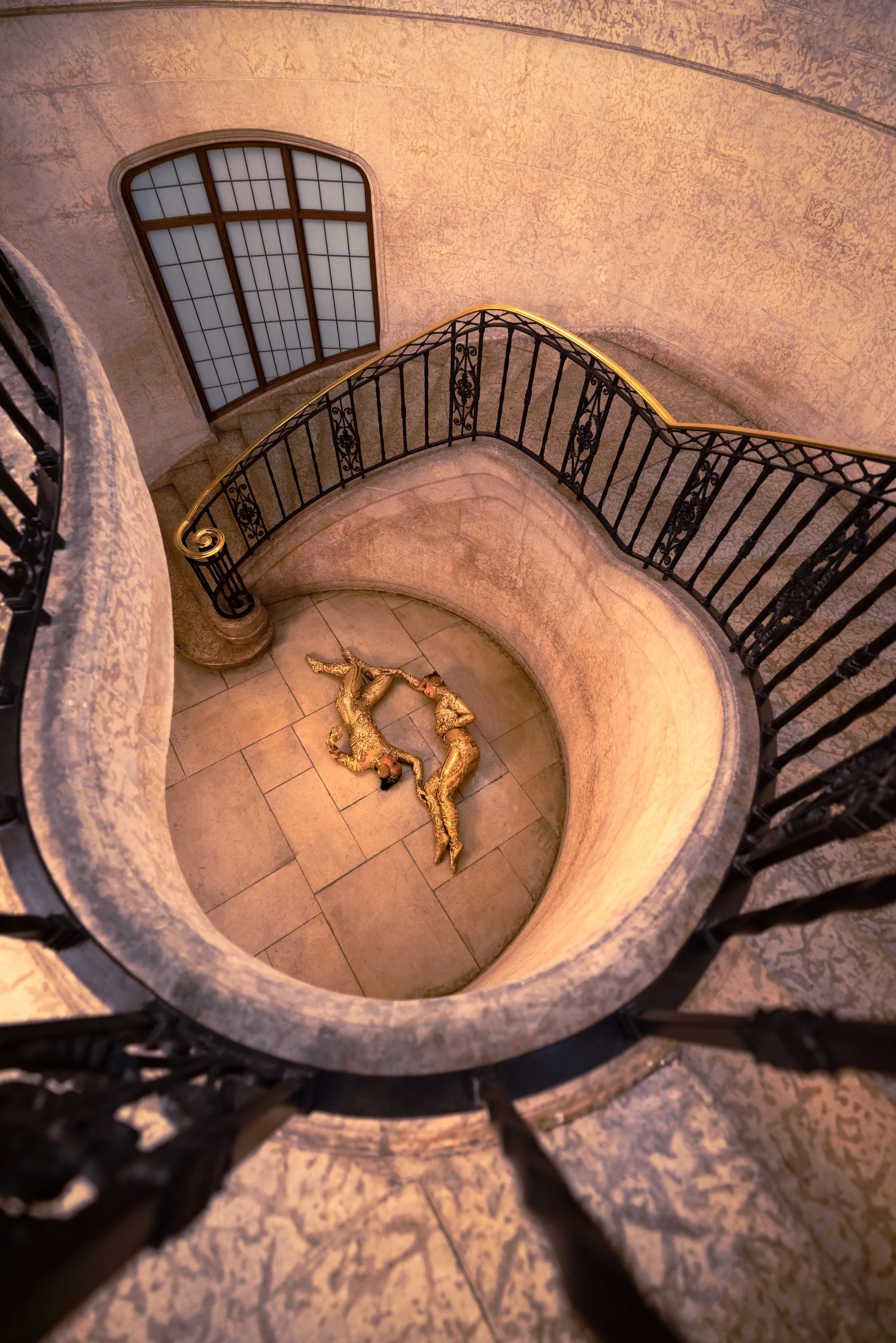 Looking down at two dancers in gold costumes lying on a tiled floor at the bottom of a staircase with black wrought-iron railing, inside a building with an arched window.