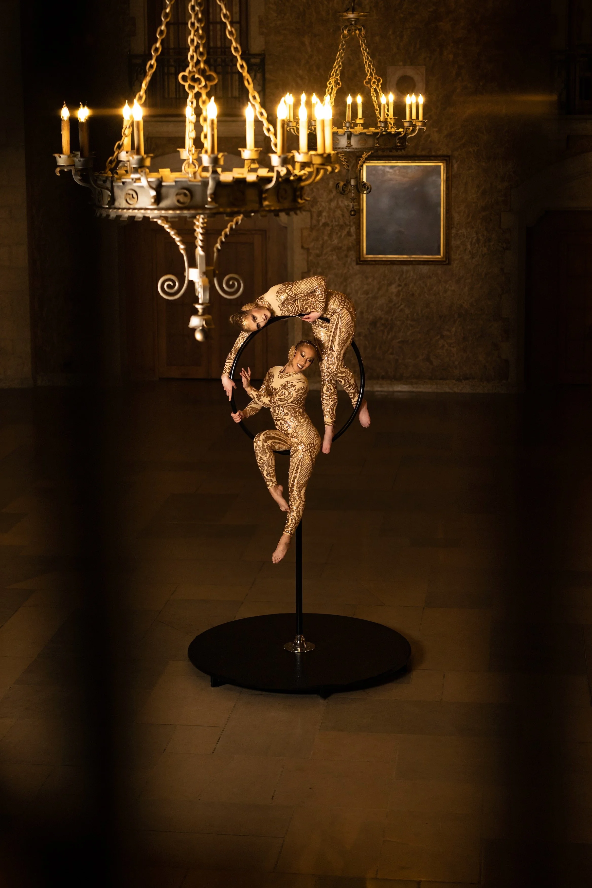 Two female aerial performers dressed in gold costumes performing on a black aerial hoop with a chandelier hanging above them in a dimly lit room.