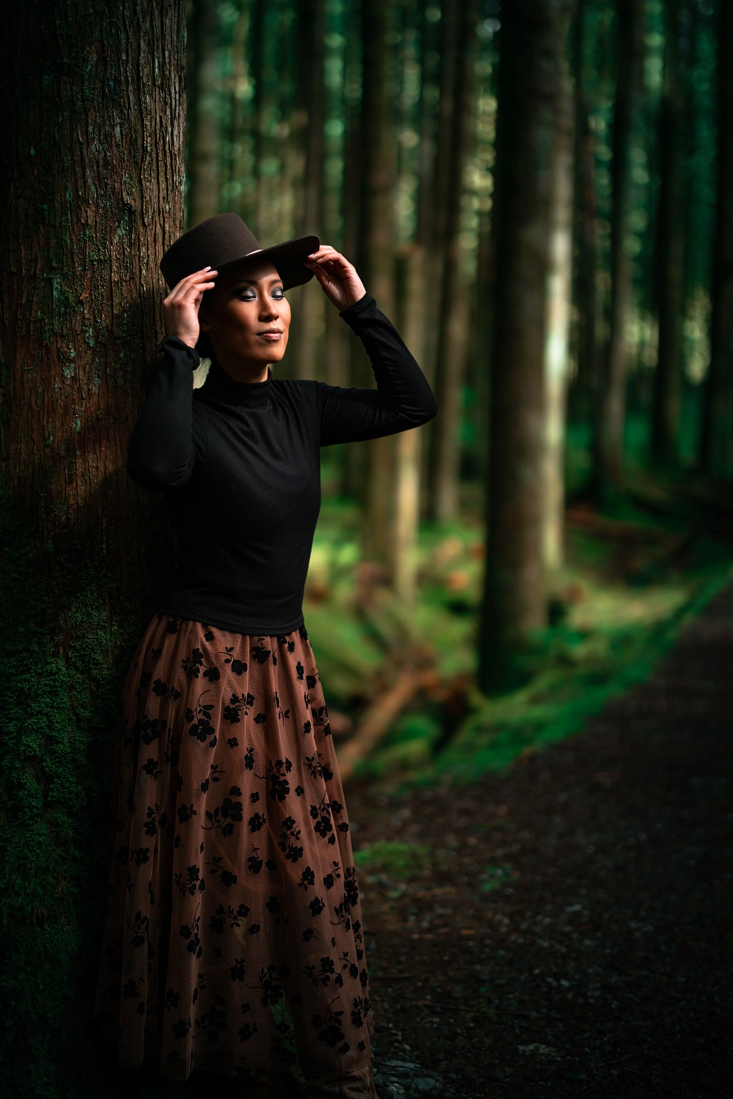 A woman with dark hair, wearing a black long-sleeve top, a brown skirt with black floral patterns, and a brown hat, stands in a dense forest leaning against a tree.