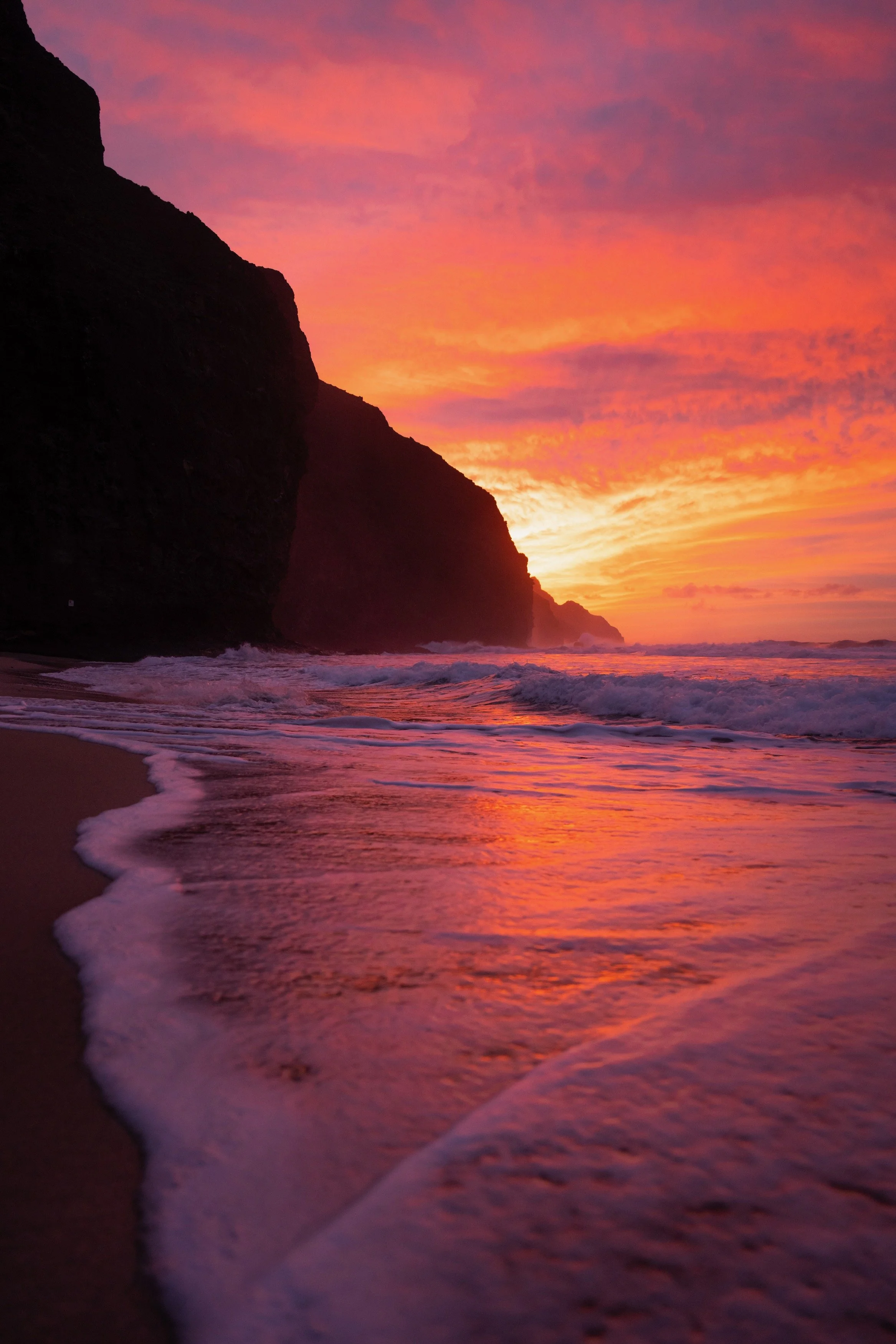 Sunset over the ocean with colorful sky, silhouetted cliffs, and gentle waves on the shore.