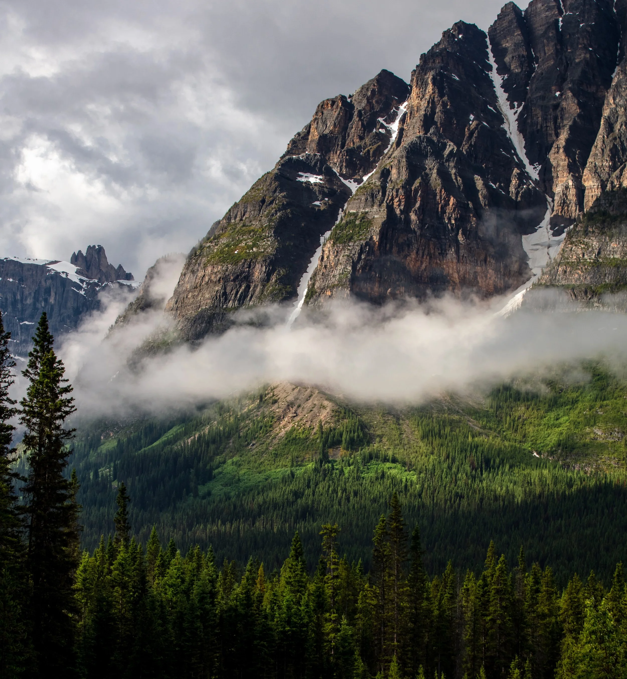 Majestic mountain landscape with snow patches, layered clouds, and dense green forest at the base.