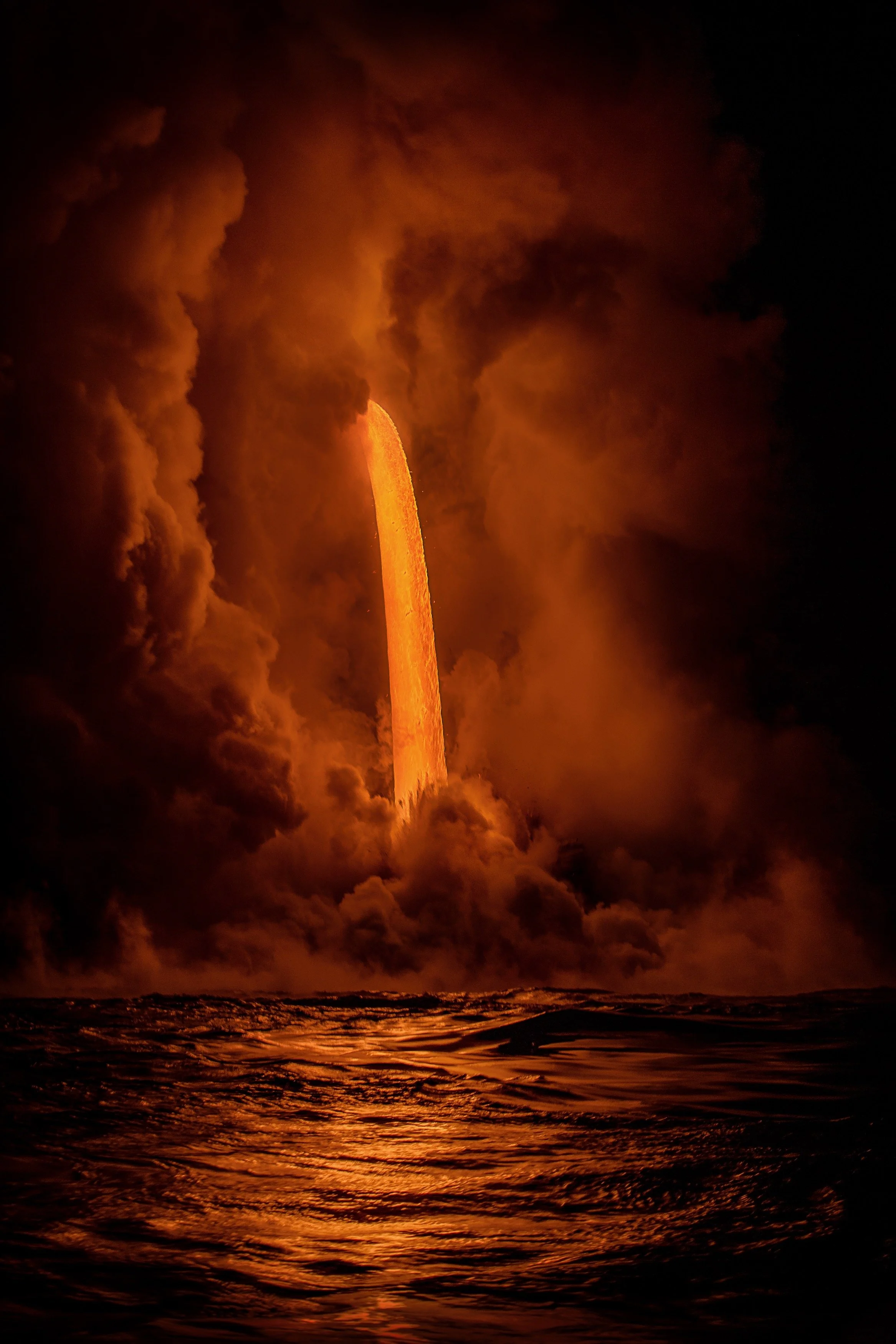 A large volcanic eruption with a fiery plume rising from the ocean at night, illuminated by lava.