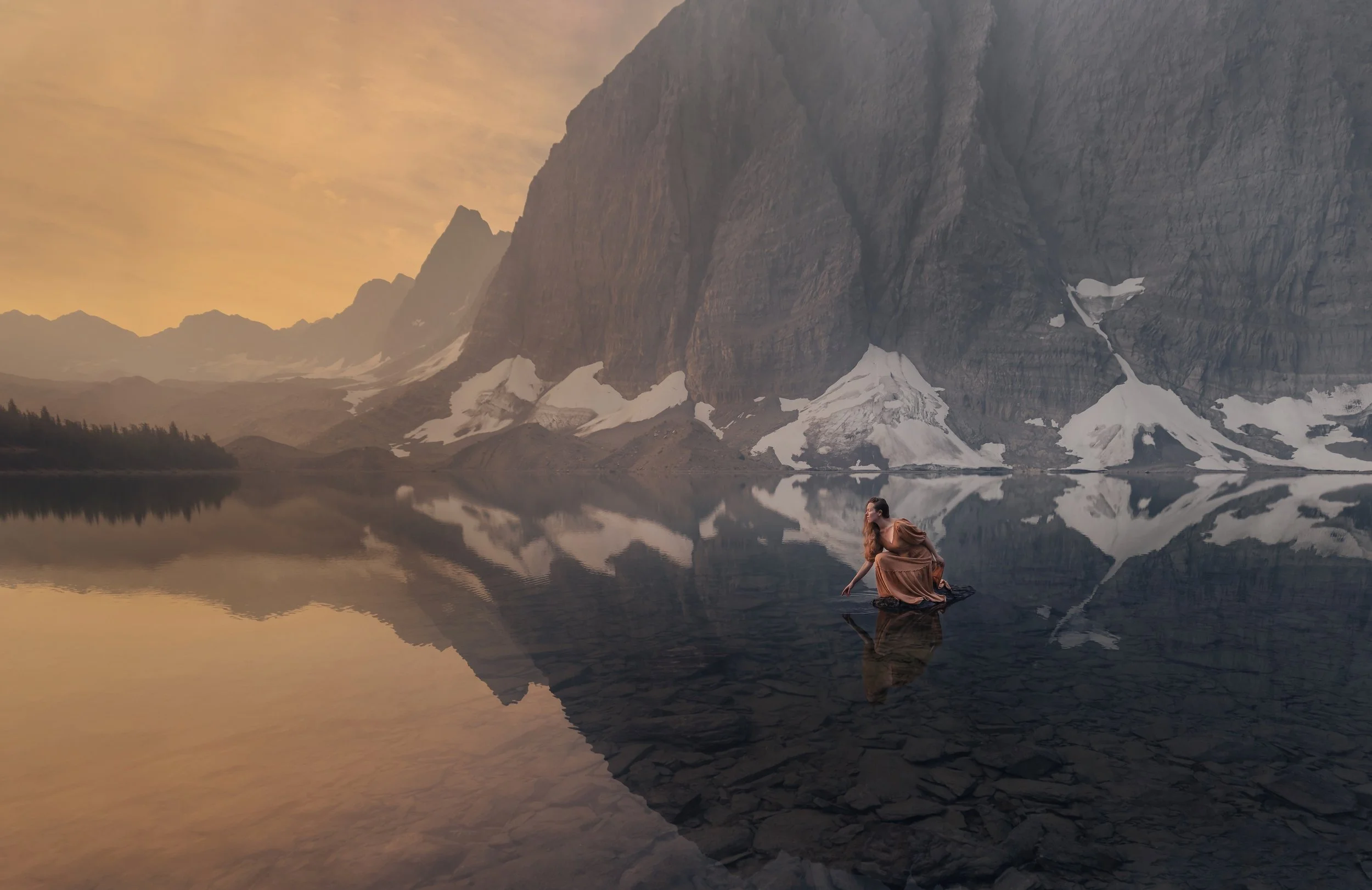 A woman in a brown dress kneeling at the edge of a calm mountain lake, with mountains and snowy patches reflected in the water during sunset.