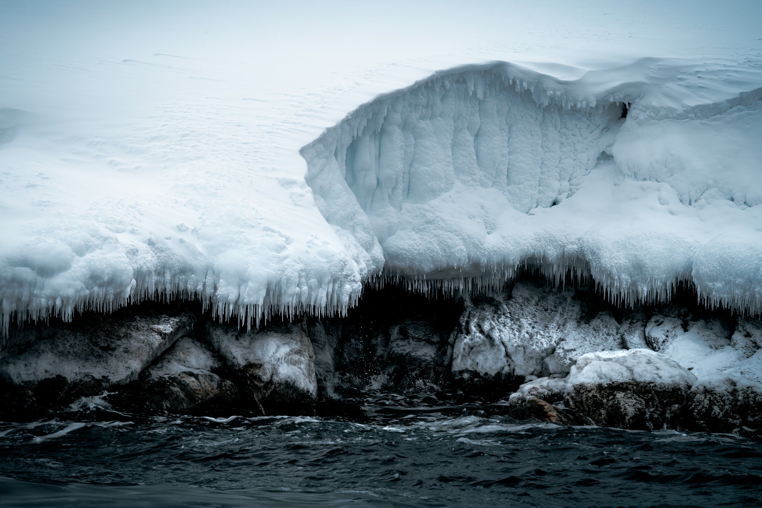 Iceberg with icicles and snow over rocks in cold water.