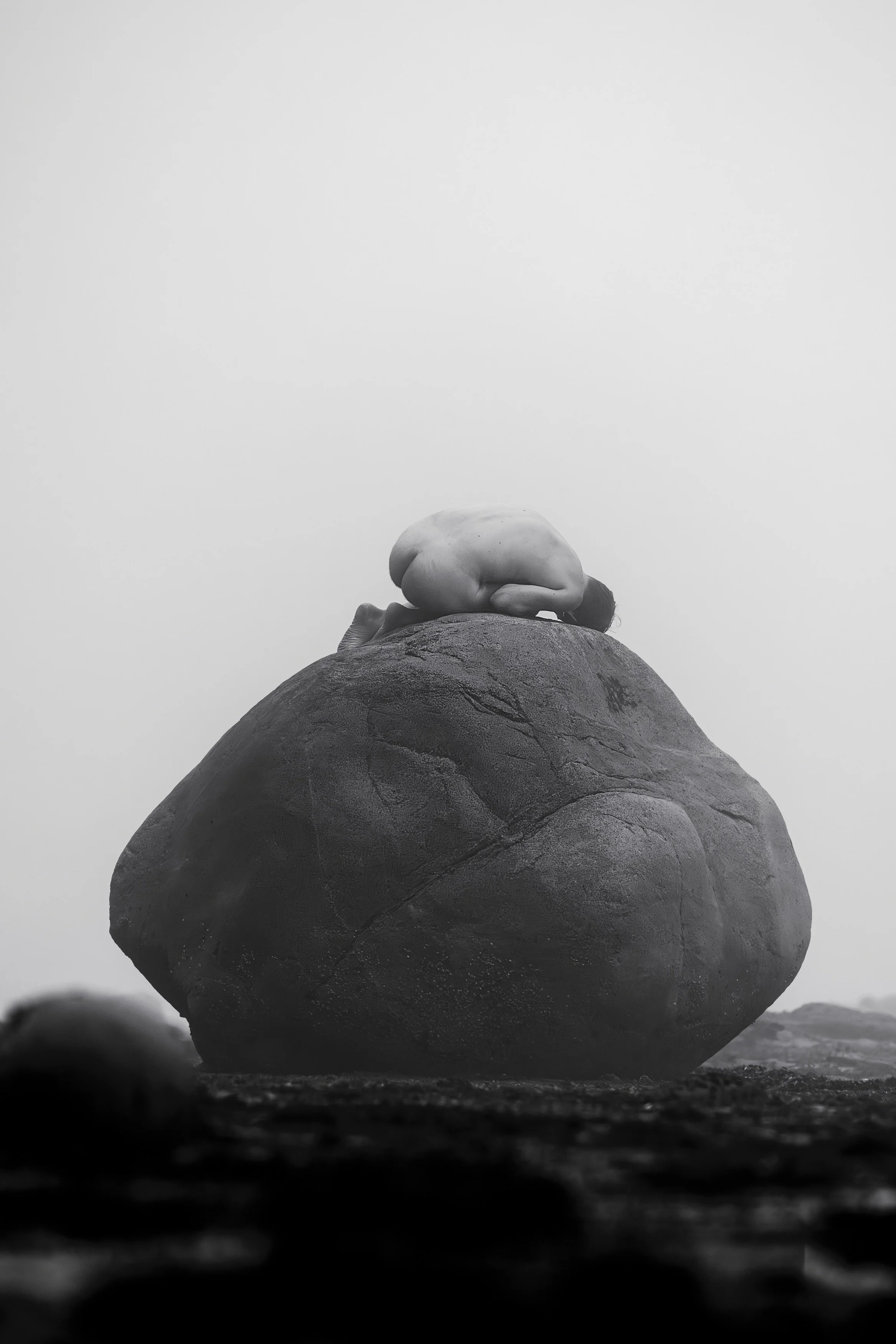 A person crouched on top of a large rock, viewed from below, in a black-and-white photograph.