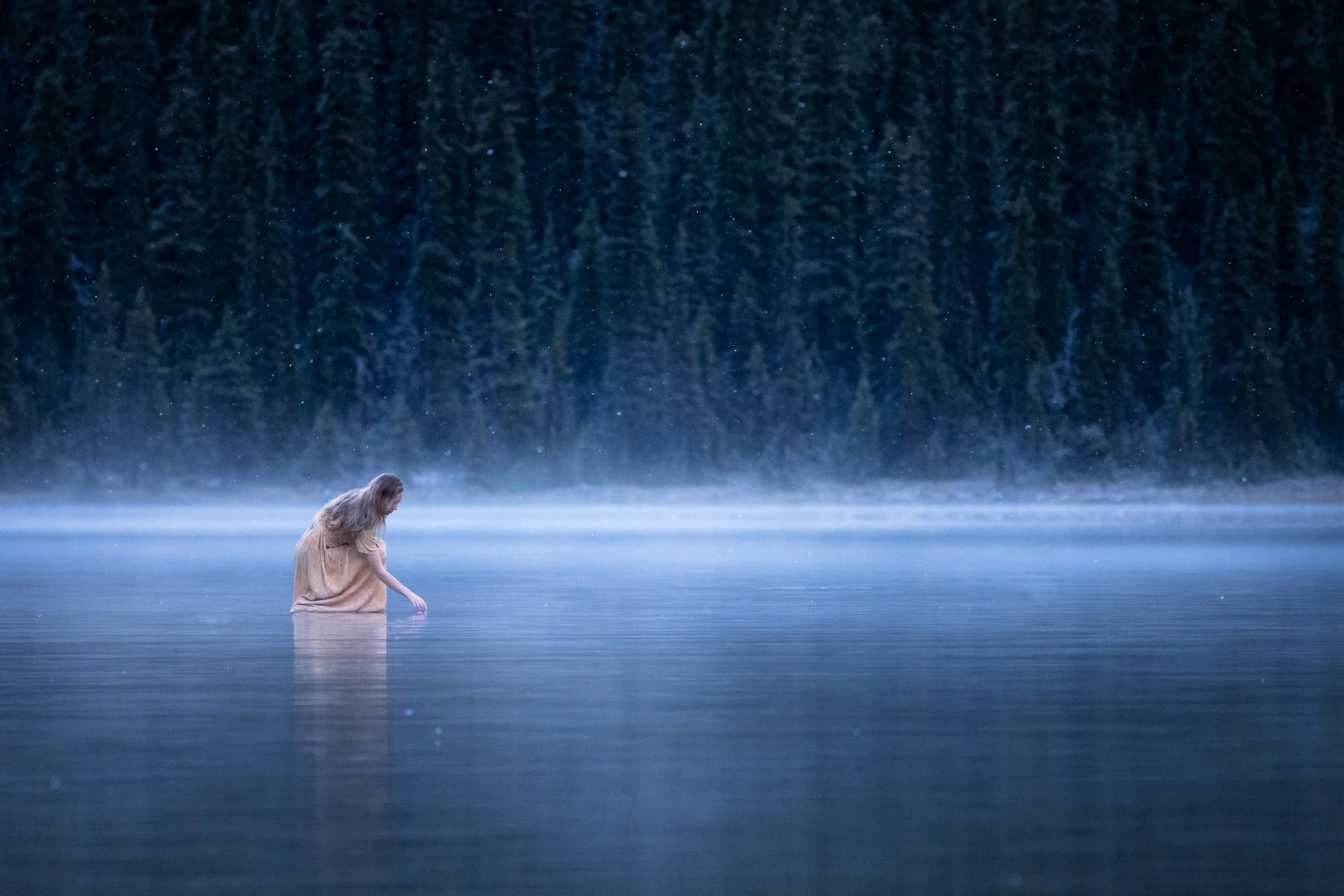 A woman in a beige dress standing in a calm body of water at dusk, with a foggy shoreline and dense forest of evergreen trees in the background.