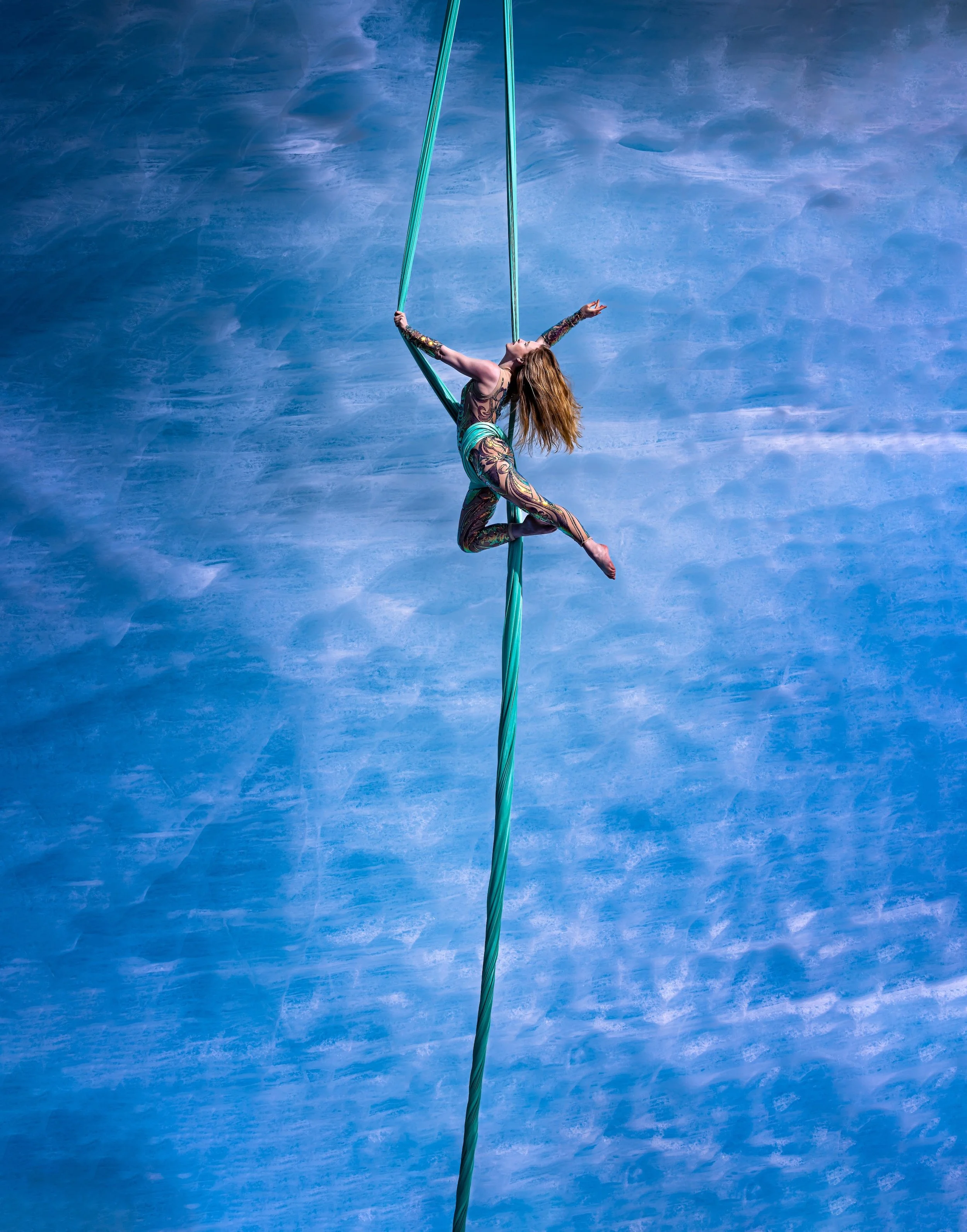 A female aerialist performing on aerial silk against a blue ice-cave wall background, wearing a colorful, patterned costume.