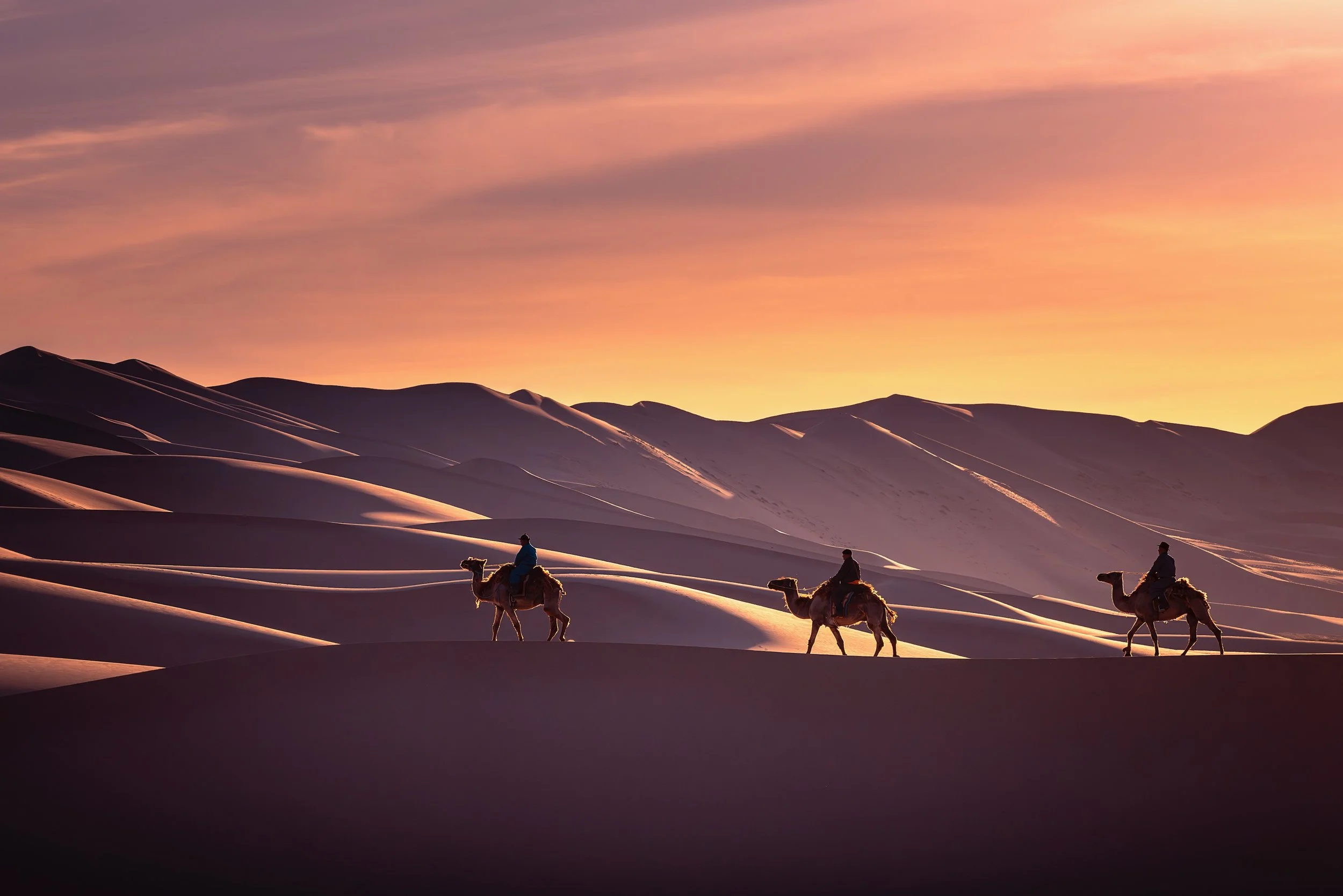 Three people riding camels across sand dunes during sunset with a colorful sky.