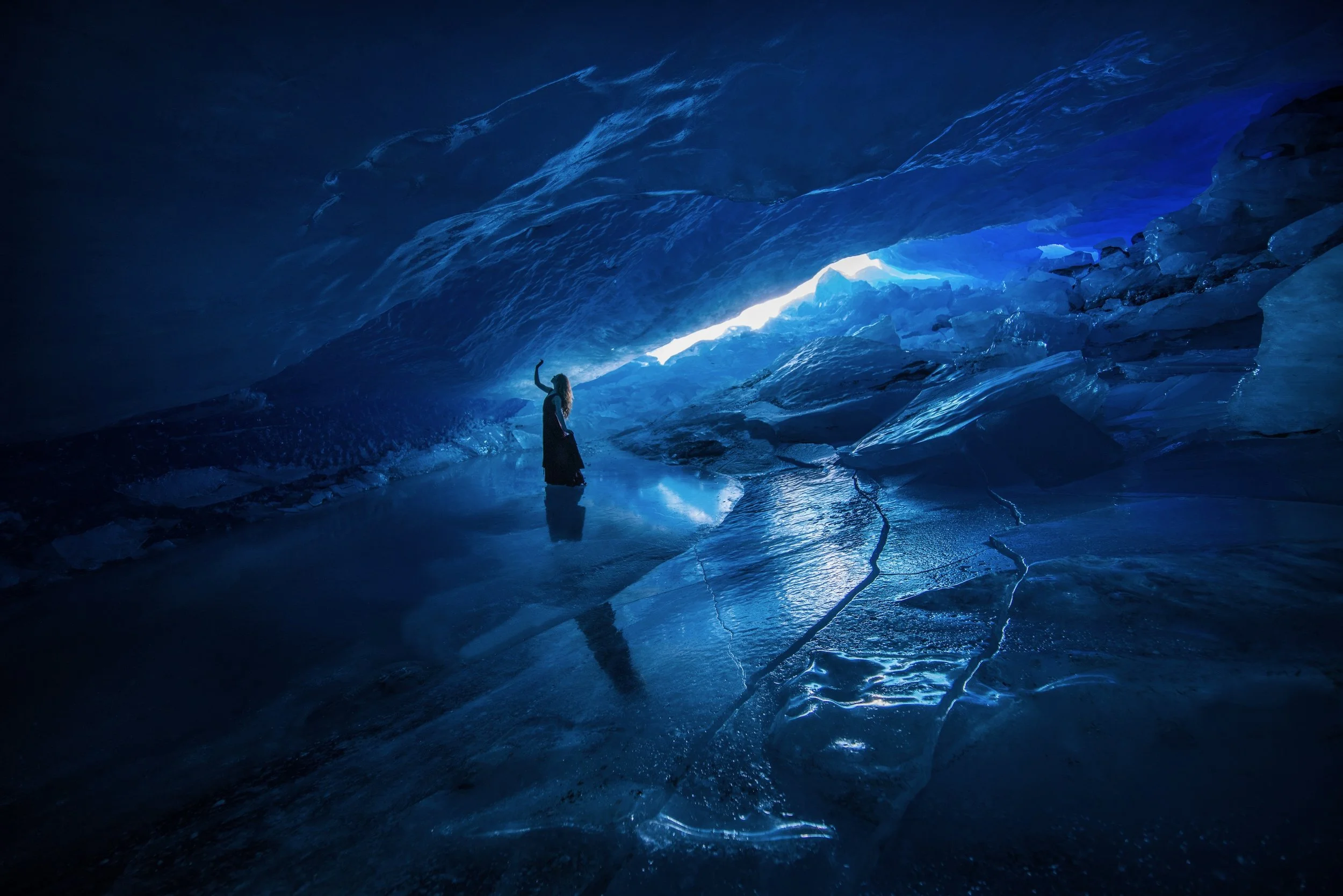 A person stands inside an ice cave with blue icy walls, illuminated by natural light coming from the entrance, and a reflective icy floor.