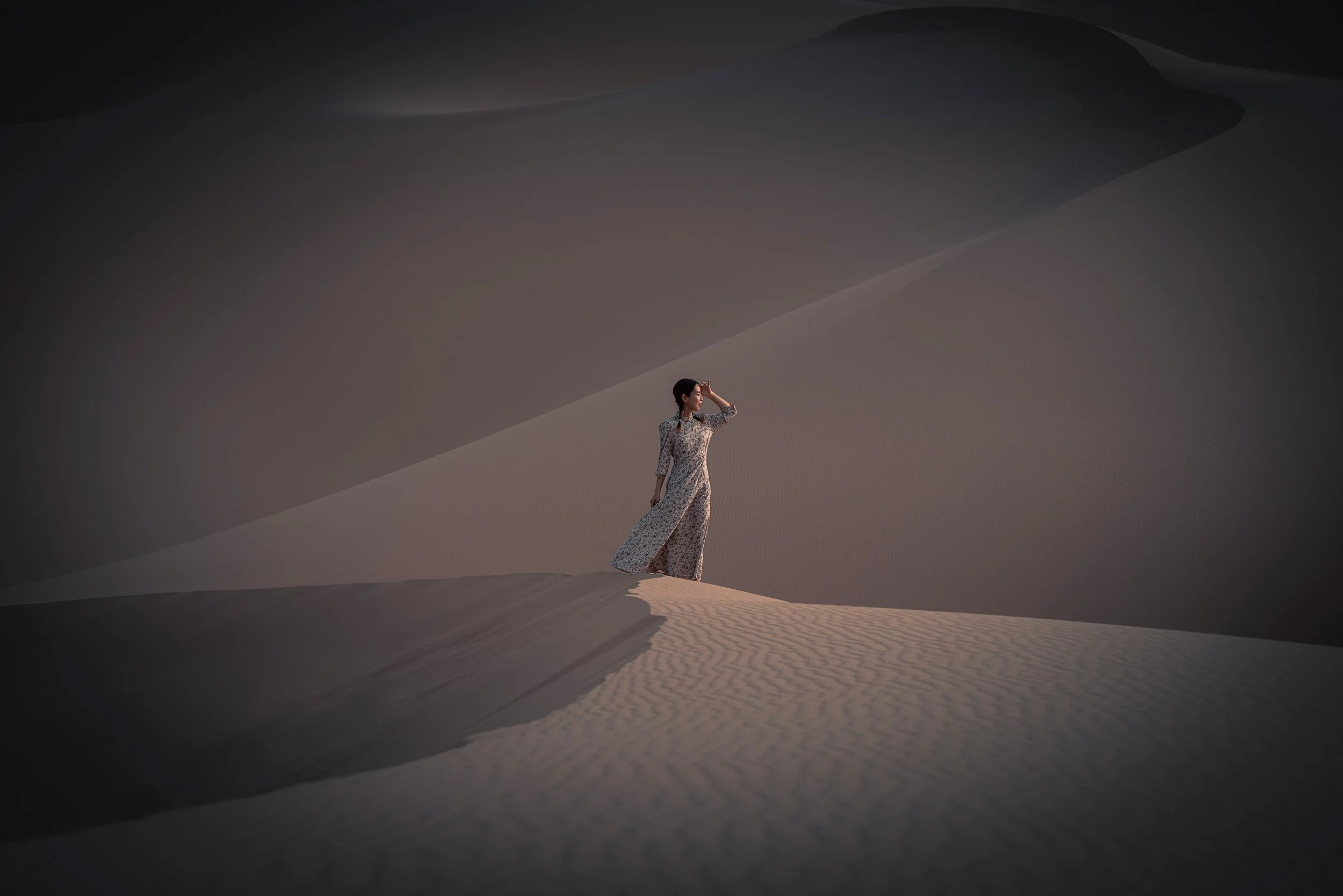A woman standing on a sand dune in a desert, wearing a long patterned dress and shielding her eyes from the sun.