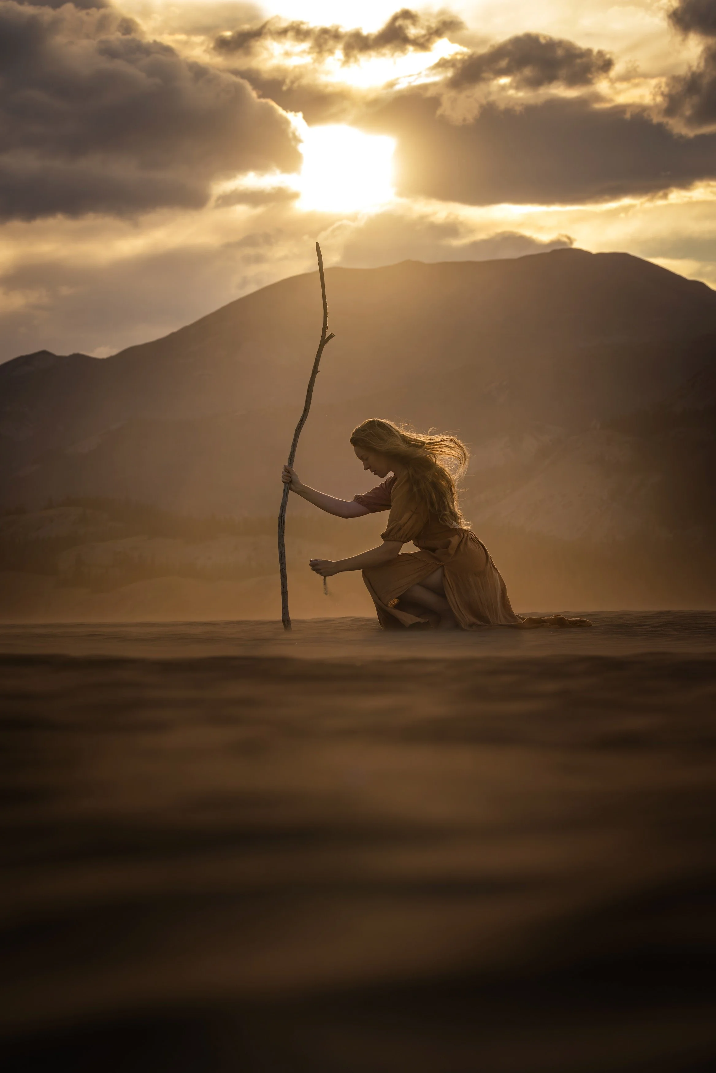 A woman kneeling in shallow water at sunset, holding a tall, thin stick, with mountains and a partly cloudy sky in the background.