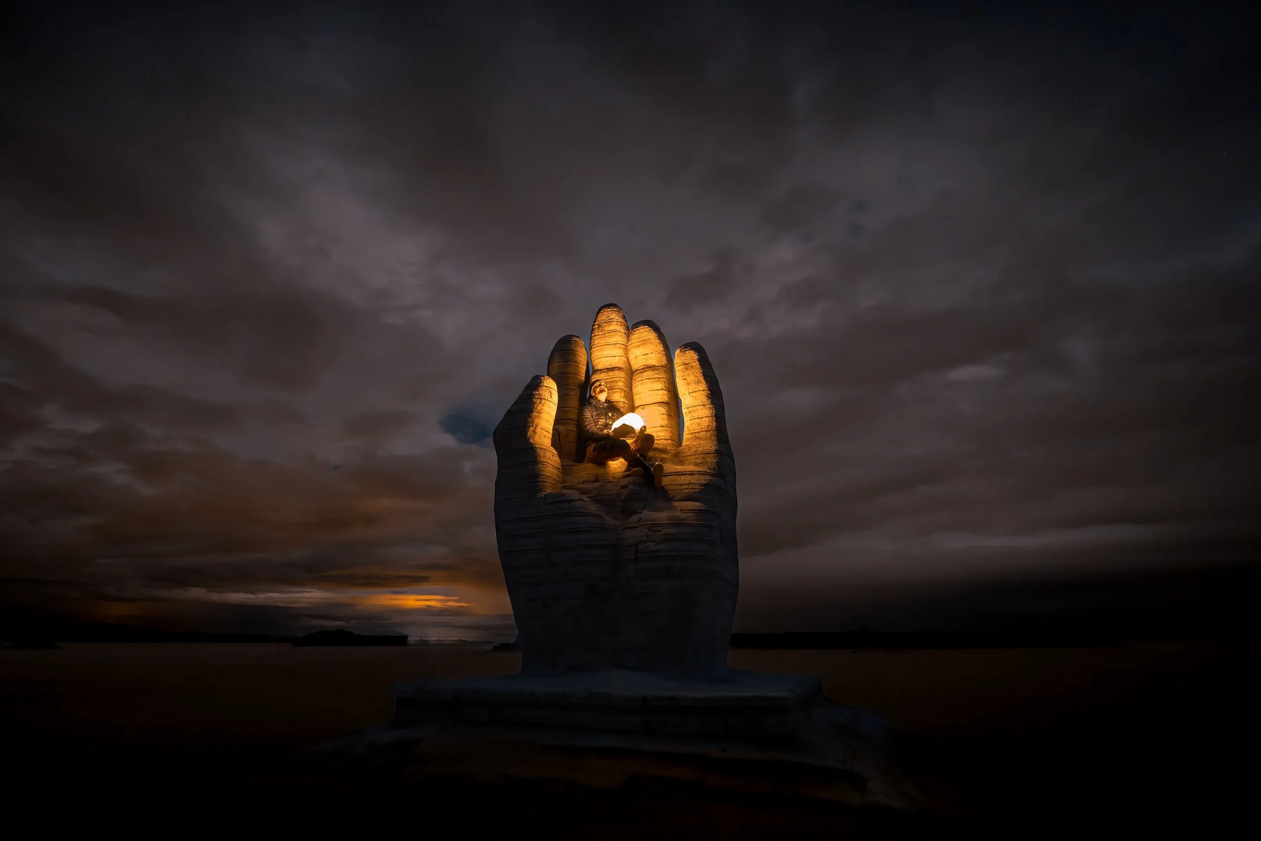 A large sculpture of a hand holding a person, illuminated against a dark, cloudy sky at dusk or night.