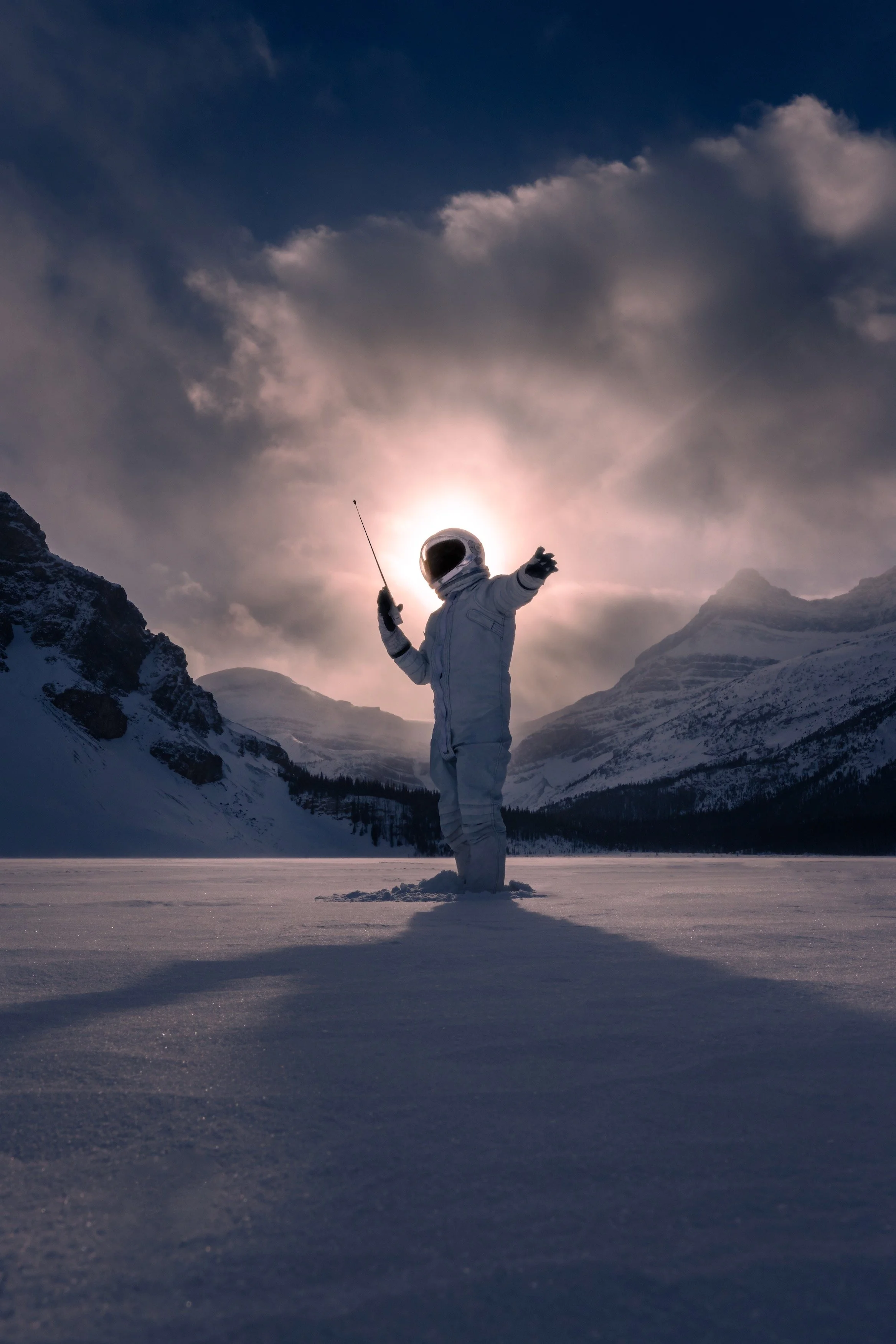 An astronaut in a white spacesuit standing in snow-covered landscape, holding a radio in one hand, with mountains and cloudy sky in the background.