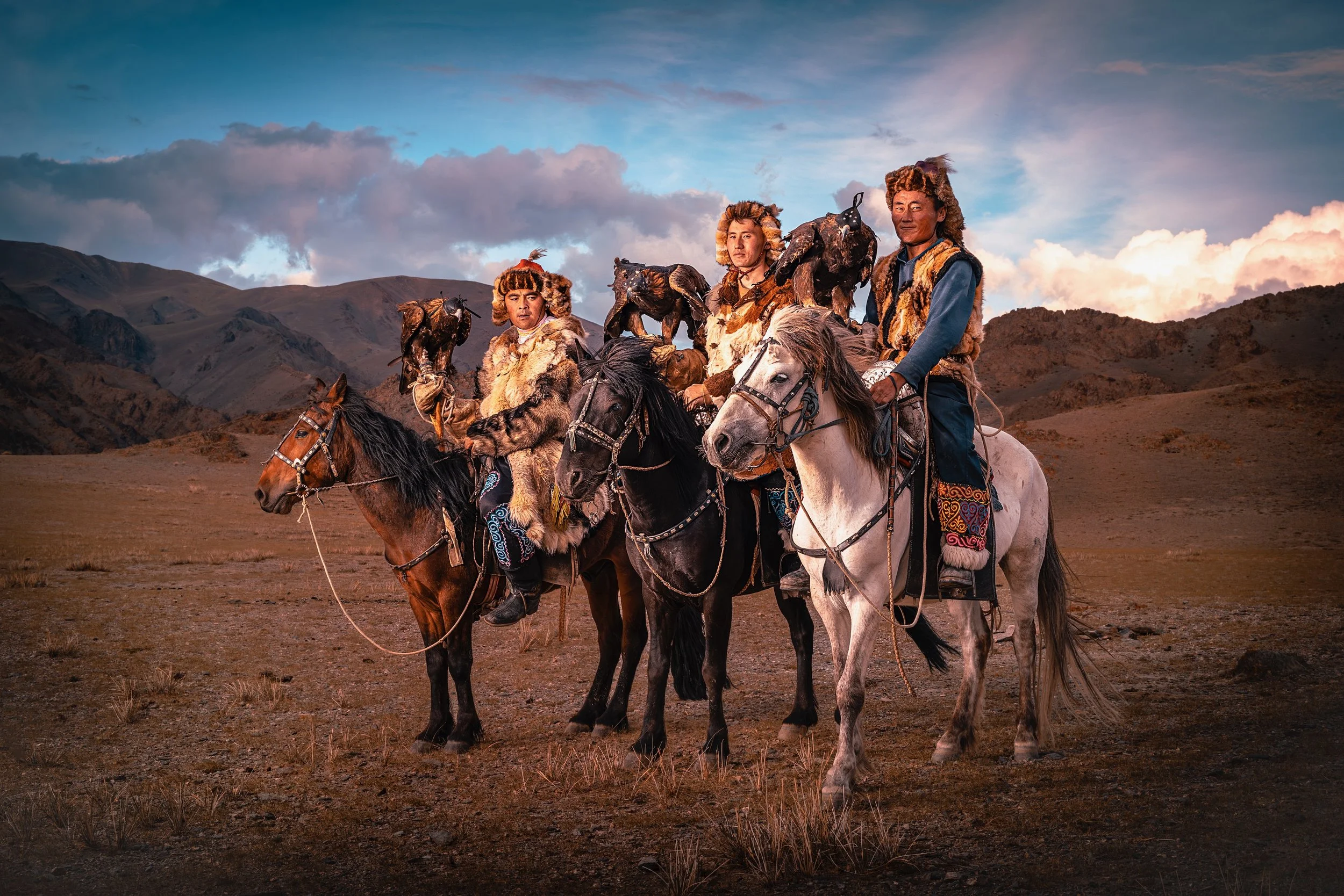 Four Mongolians dressed in traditional costumes on horseback, holding eagles, in a mountainous landscape during sunset.