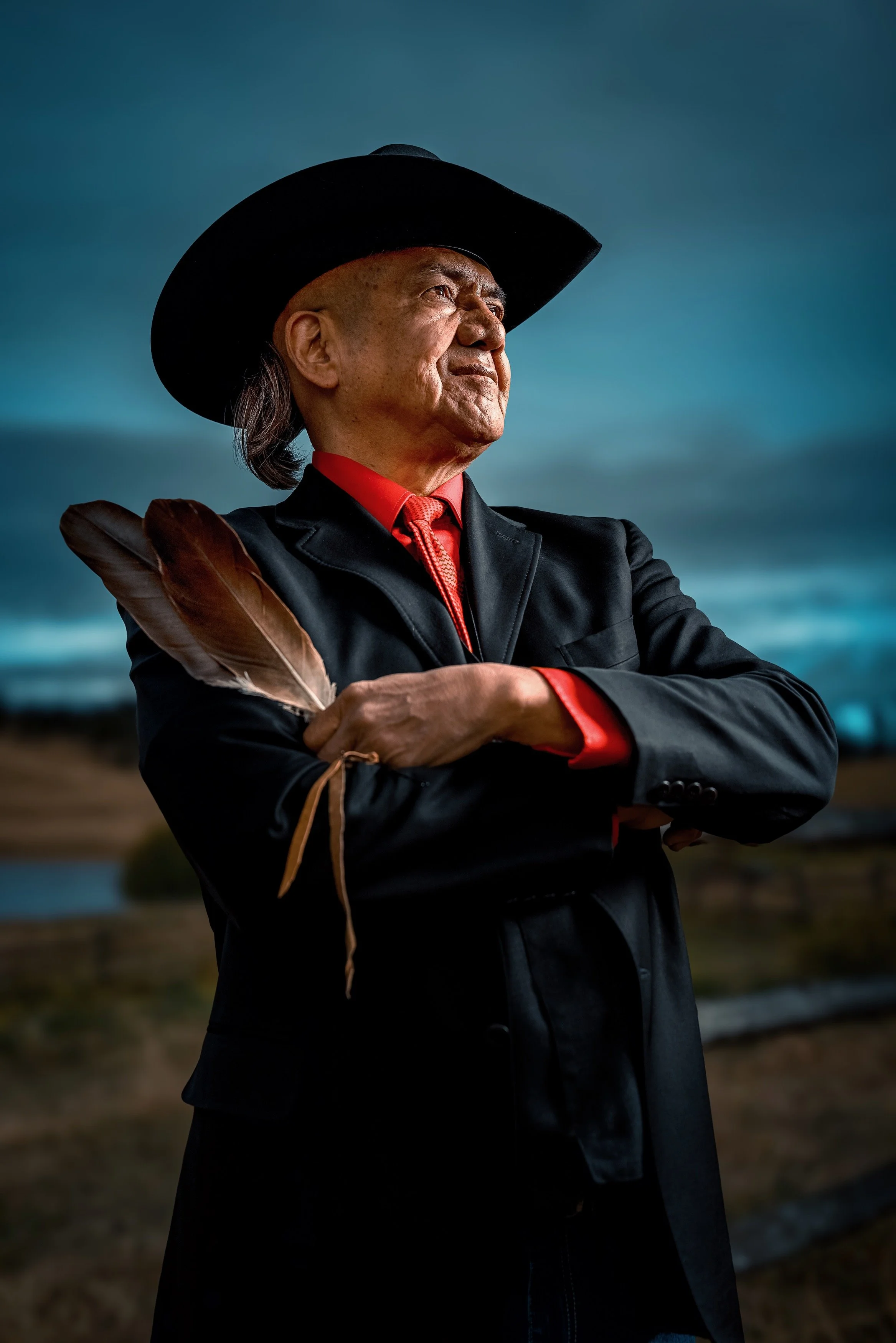 An elderly Indiginous man standing outdoors against a cloudy sky, wearing a black cowboy hat, a black jacket, and a red shirt with red accents, holding a feather in his hand.