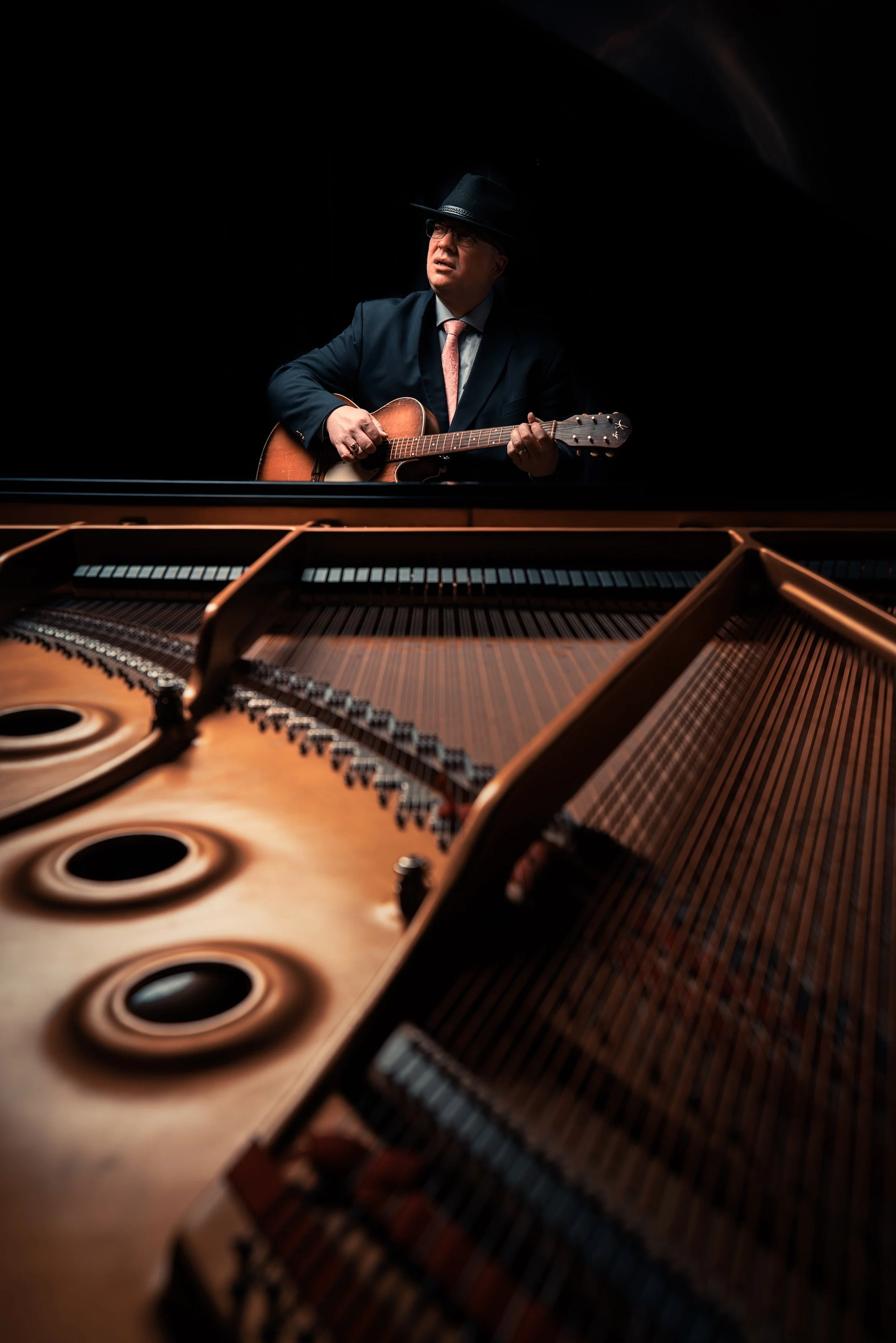 A man wearing a suit, fedora, and glasses plays an acoustic guitar behind a grand piano in a dark setting.