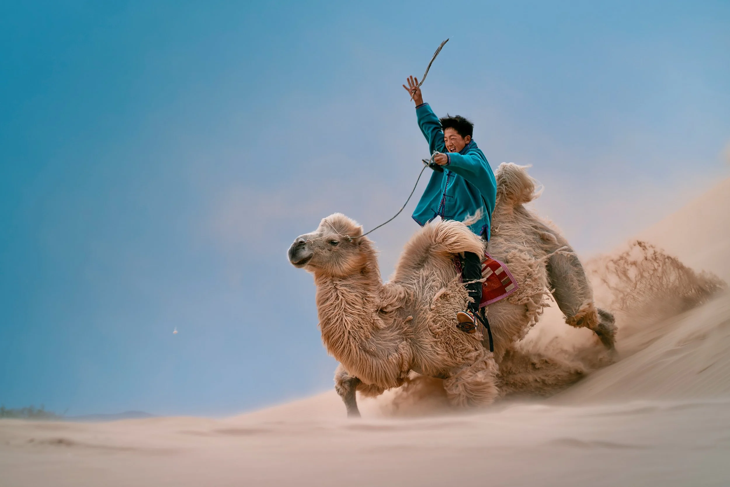 Person riding a camel in the desert, smiling and holding a whip, with sand and a clear blue sky in the background.
