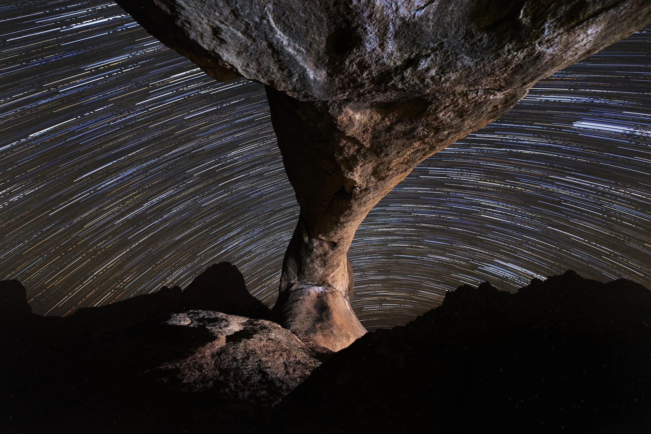 Long-exposure photograph of star trails in the night sky viewed from beneath a large rock formation with a curved, balanced rock shape.