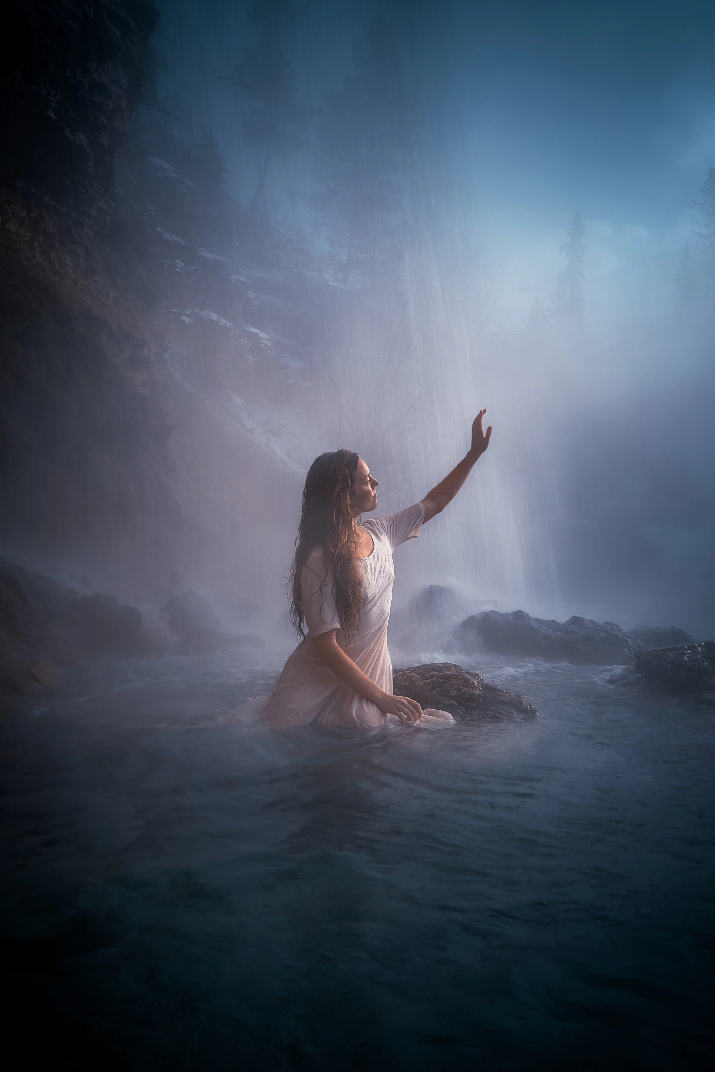 A woman in a white dress kneels in a river with a waterfall in the background.