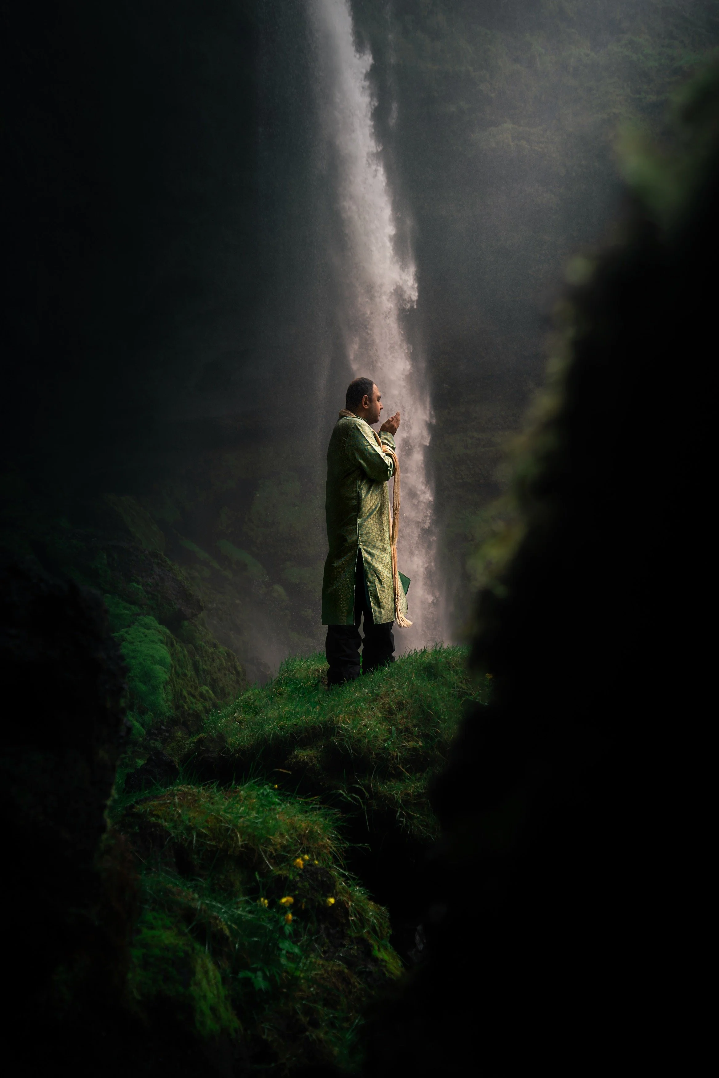 A man dressed in a traditional robe standing on a grassy mound near a waterfall, seen through a natural frame of dark rocks or foliage.