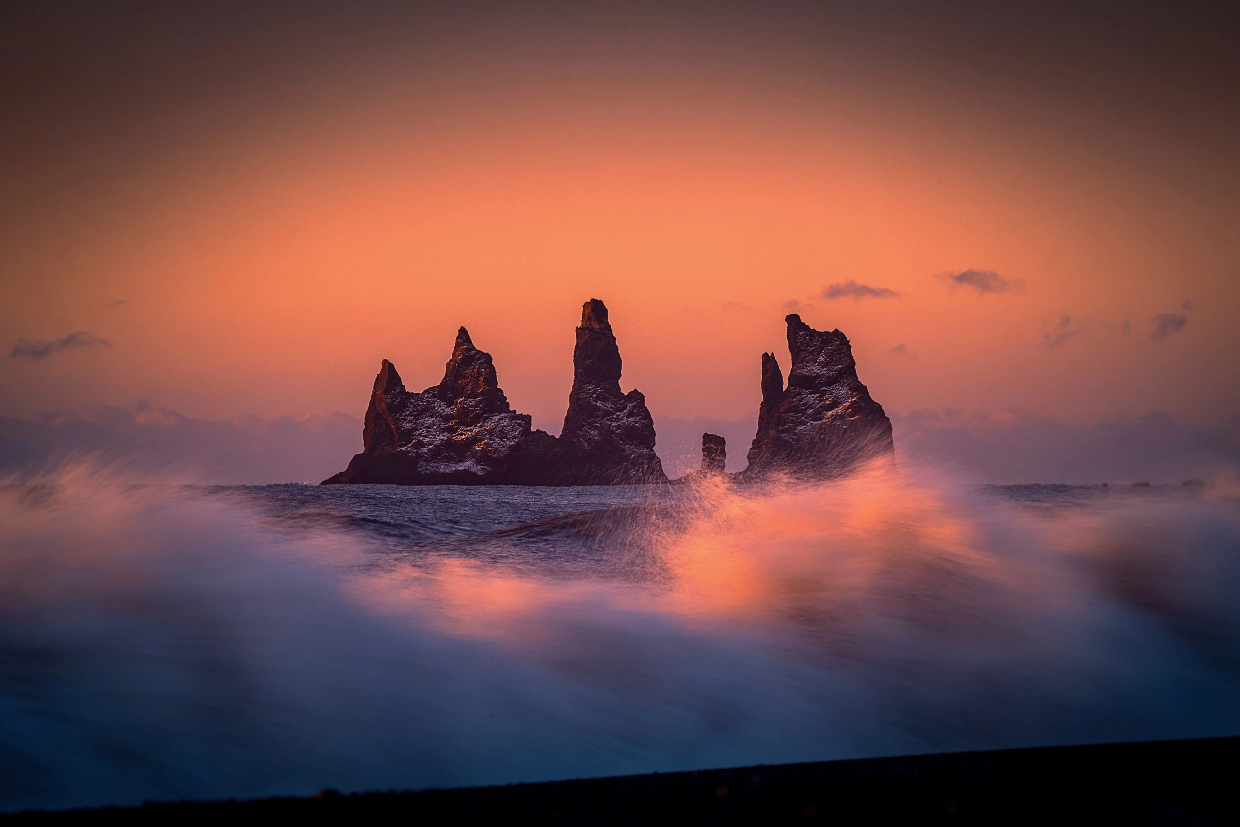 Rock formations rising from the ocean during sunset with orange and pink sky and waves crashing.