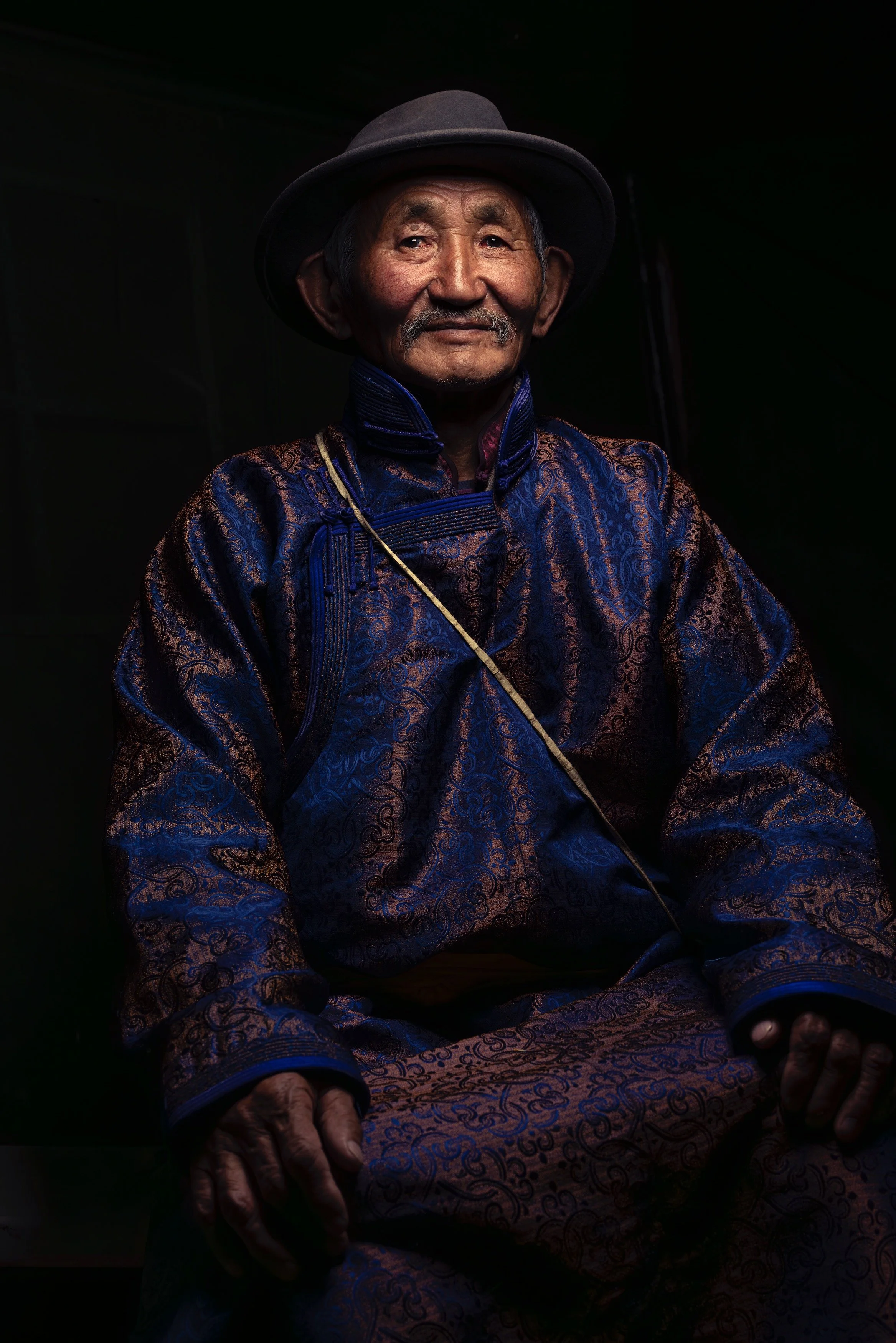 An elderly man wearing traditional Mongolian clothing and a wide-brimmed hat, sitting with a calm expression.