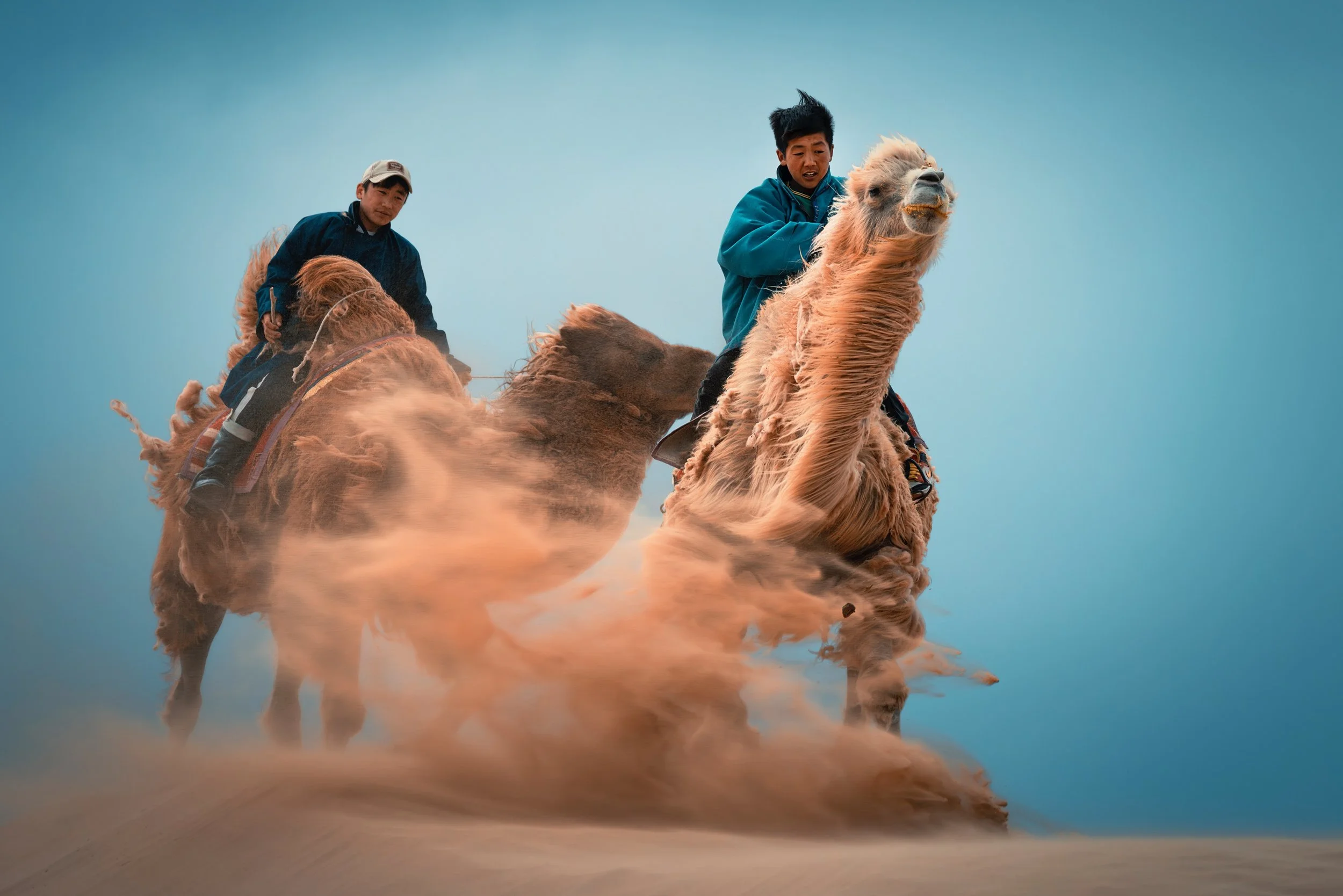 Two men riding camels through desert with dust clouds, against clear blue sky.