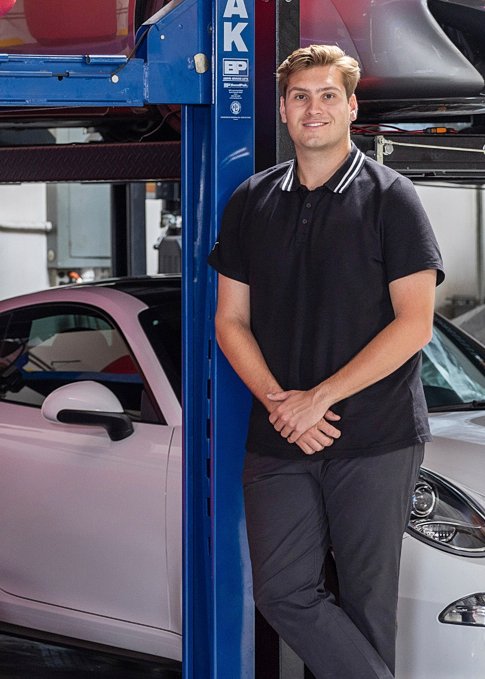 A young man with blond hair, wearing a black polo shirt and dark pants, standing with hands folded in front of him, in a car garage next to a blue car lift and two cars, one pink and one white.