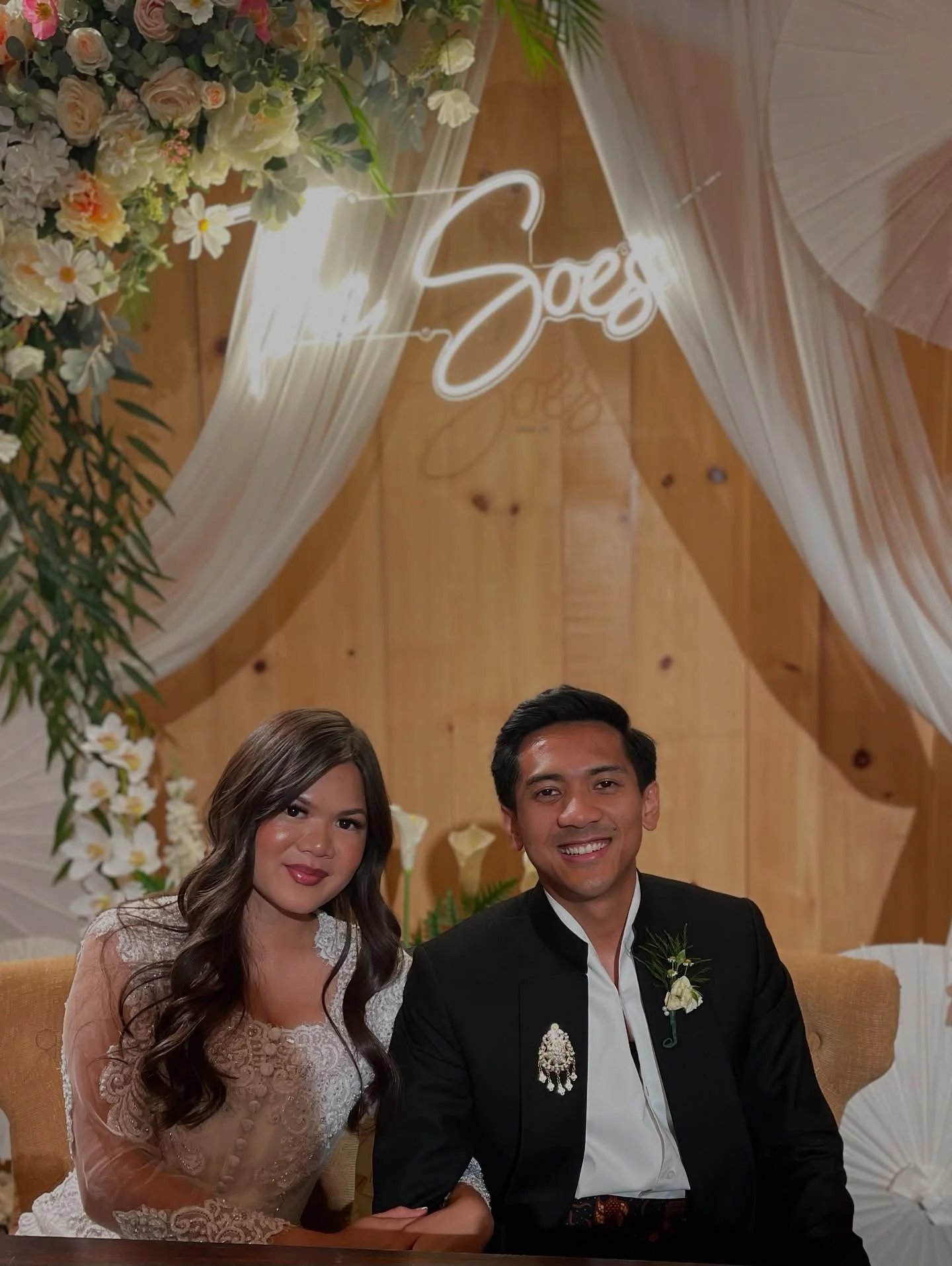 A couple sitting at a table, dressed in formal attire, in front of a decorated wooden backdrop with flowers, white drapery, and a neon sign that says 'Happily So'.