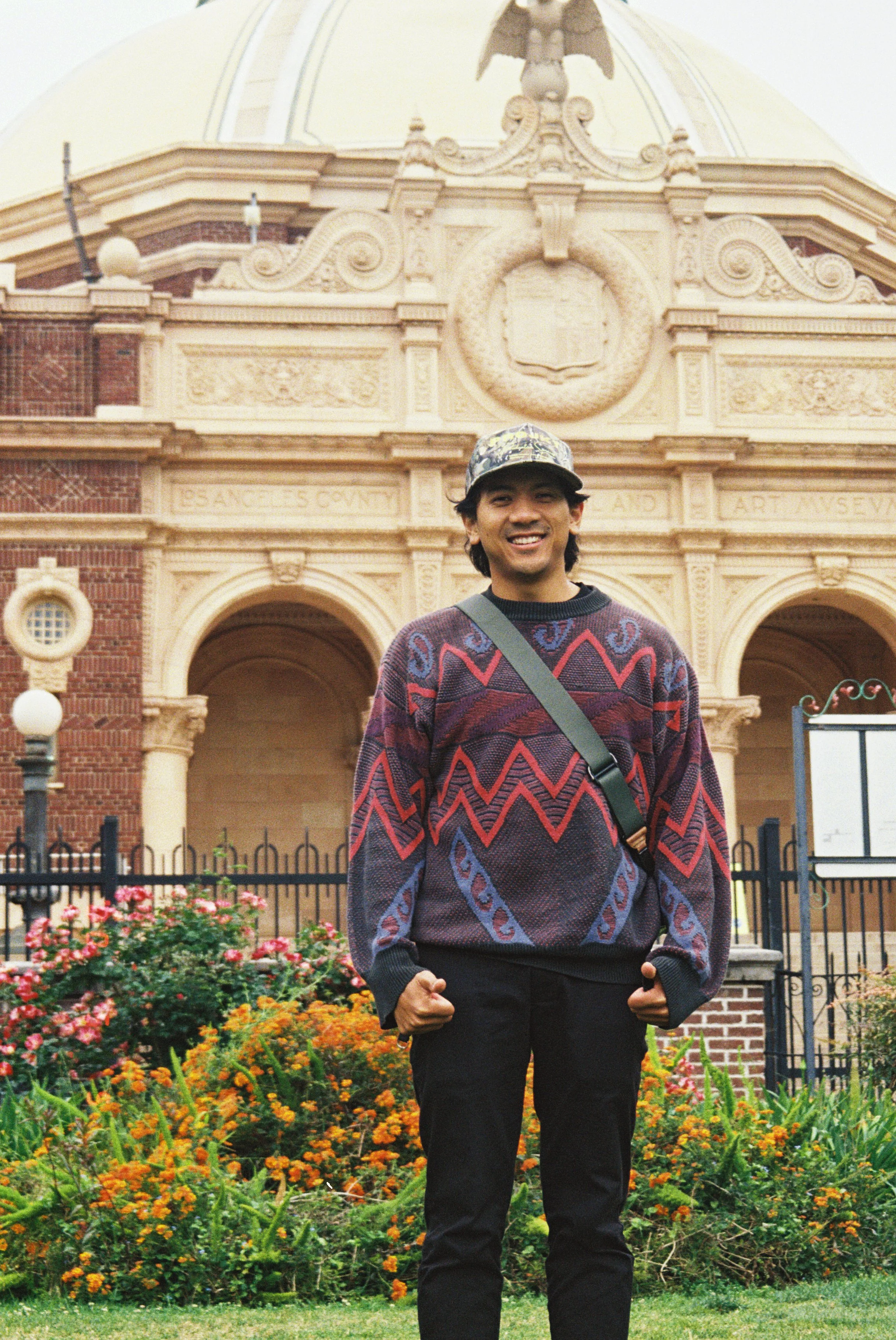 A young man wearing a colorful sweater, jeans, and a cap, standing in front of a historic building with elaborate architectural details and a garden with flowers.