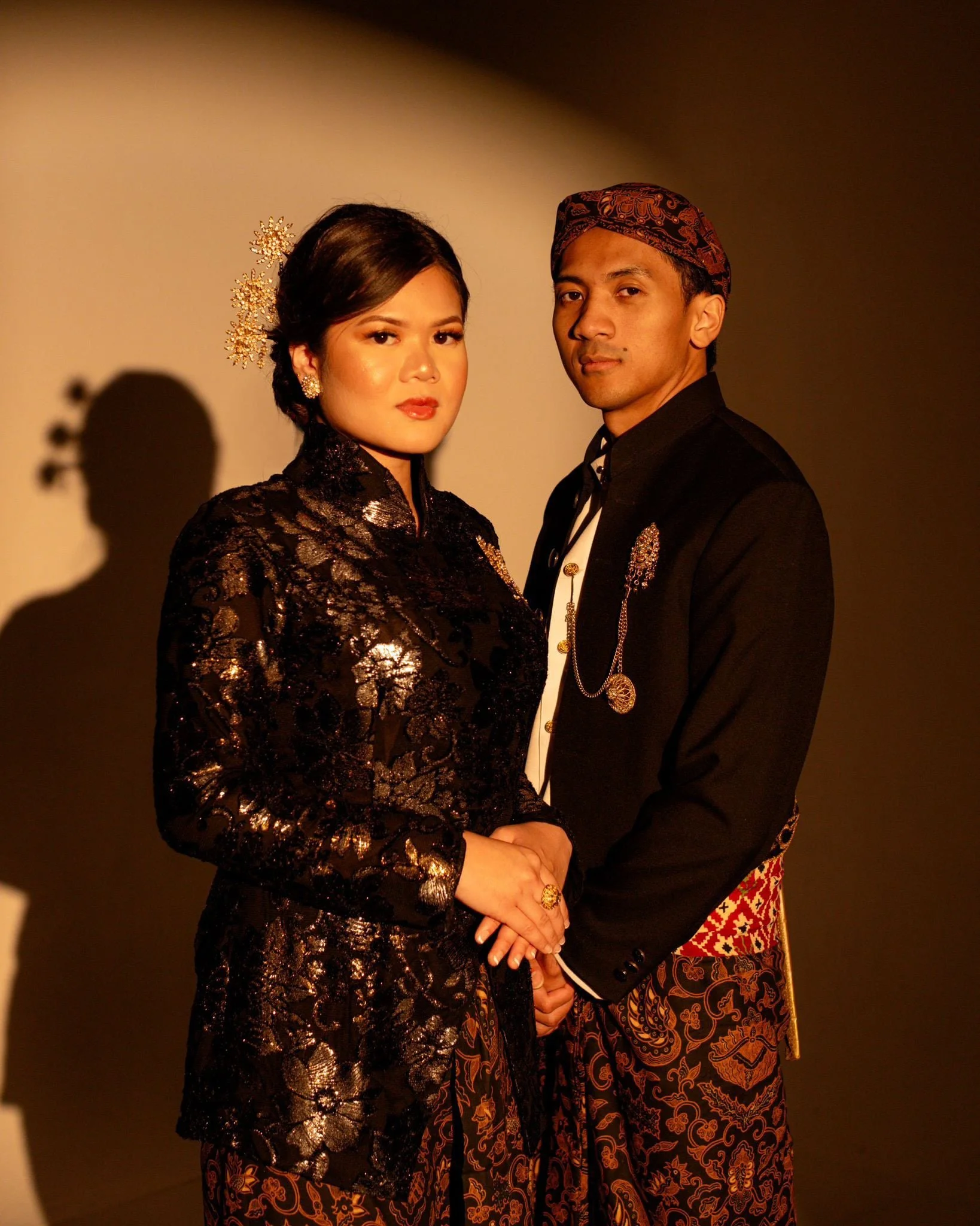 A man and woman dressed in traditional Indonesian attire standing close together against a plain background.