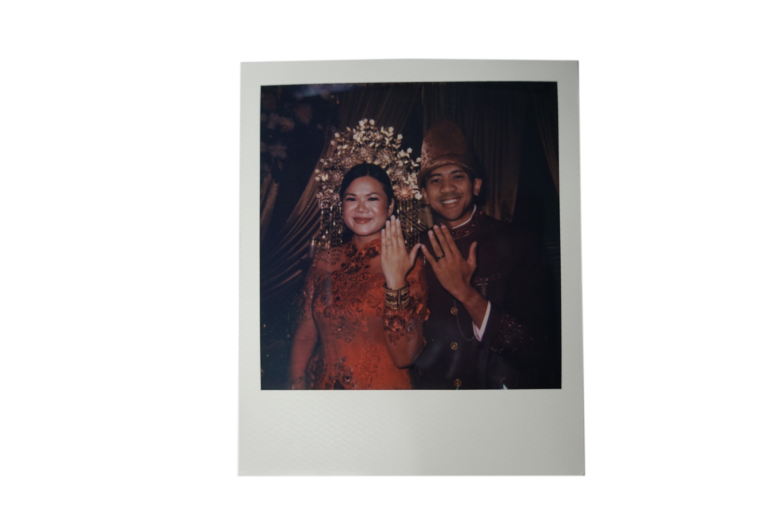 A couple in traditional Indonesian wedding attire showing off their wedding rings, standing in front of a decorated backdrop with lights and flowers.