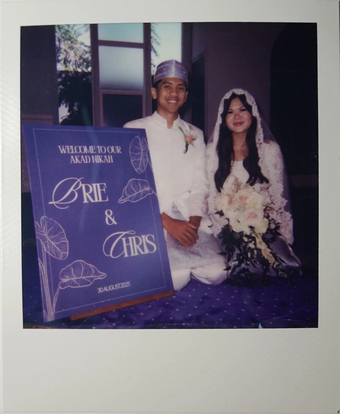 A couple dressed in traditional wedding attire sitting next to a large sign that reads, "Welcome to our akad nikah Brie & Chris, 30 August 2005." The groom is wearing a white outfit with a purple headscarf, and the bride is dressed in a white lace dress with a veil and holding a bouquet of flowers.