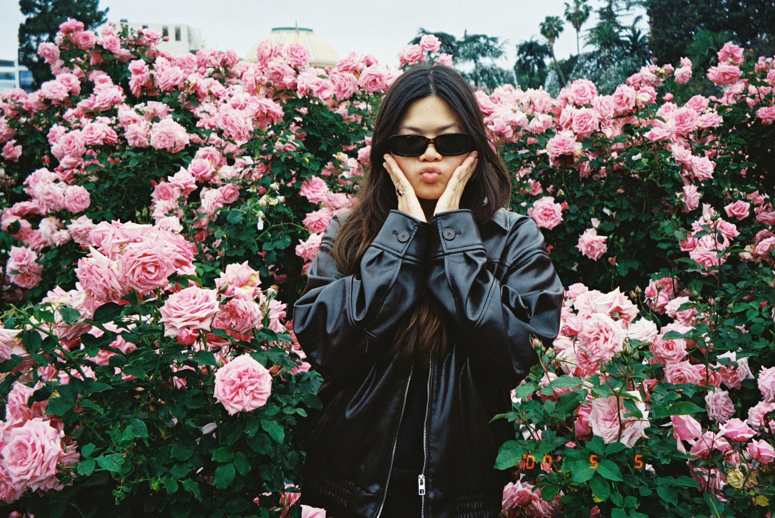 Young woman with long dark hair wearing black sunglasses and a black leather jacket standing in front of pink roses, making a kissy face with hands on cheeks.